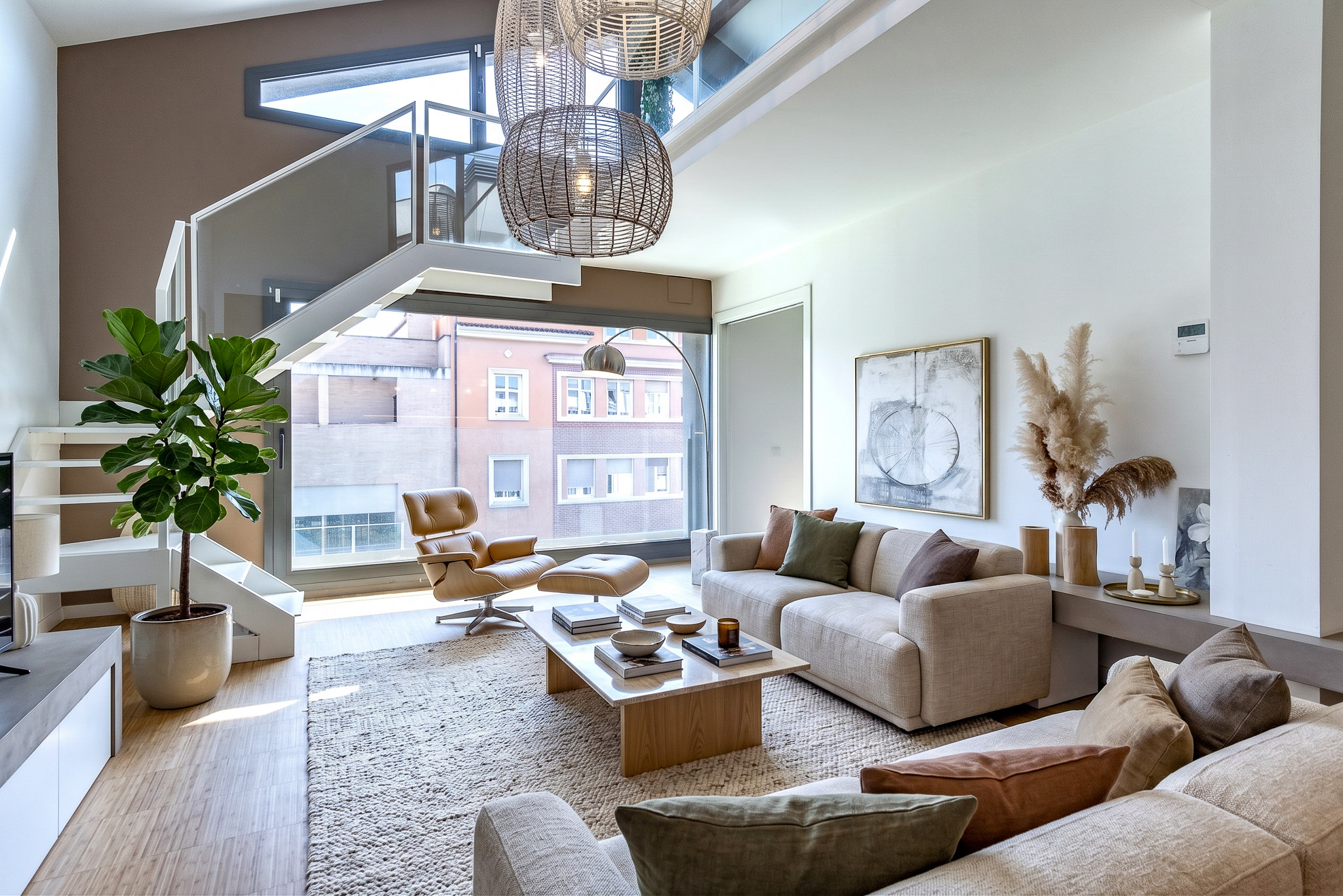 Modern living room with high ceilings, a staircase, beige sofa, lounge chair, coffee table, and large windows. [AI-generated text]