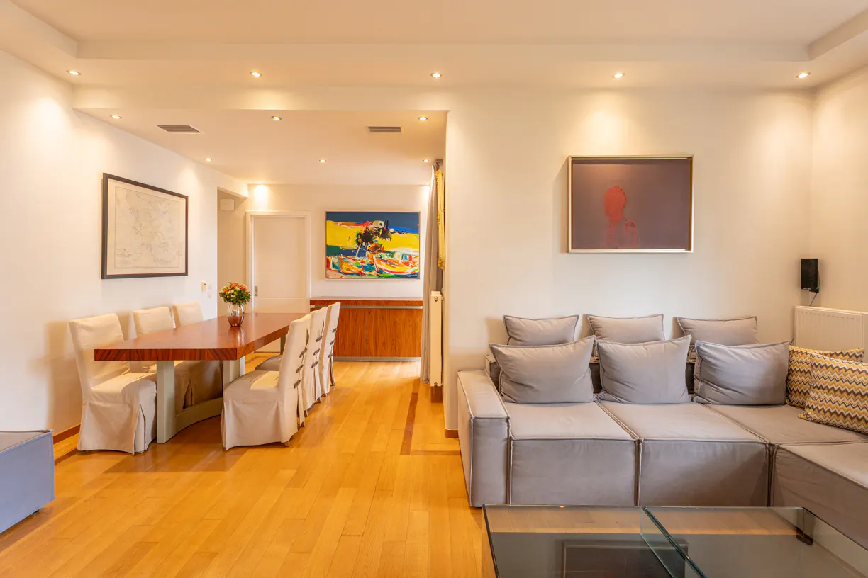 A bright living and dining area with wood floors, a gray sectional sofa, and a wood dining table with upholstered chairs. Artwork adorns the white walls.