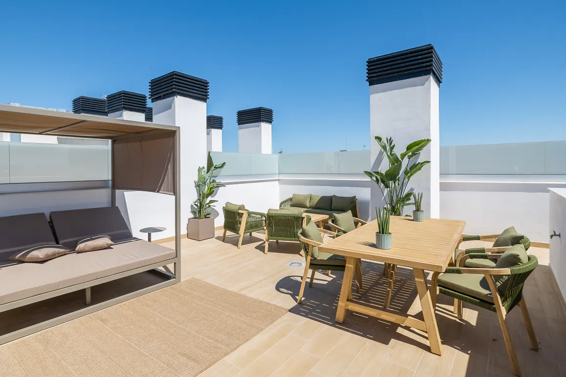 Rooftop patio with a daybed, table, and chairs. White walls and chimneys are visible against a clear blue sky. Plants add a touch of greenery.