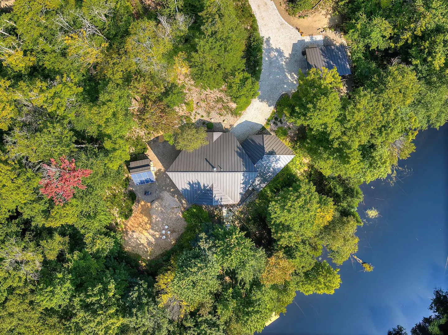 Aerial view of a modern house with a gray roof, surrounded by lush green trees and a blue lake. A gravel driveway leads to the house.