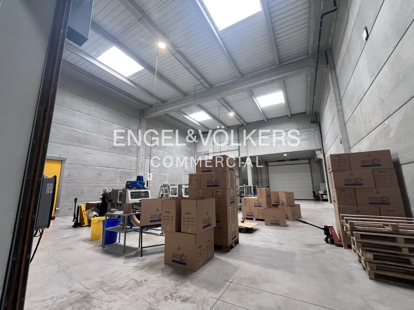 A wide shot of a warehouse interior with boxes, pallets, and equipment under a high ceiling with skylights.