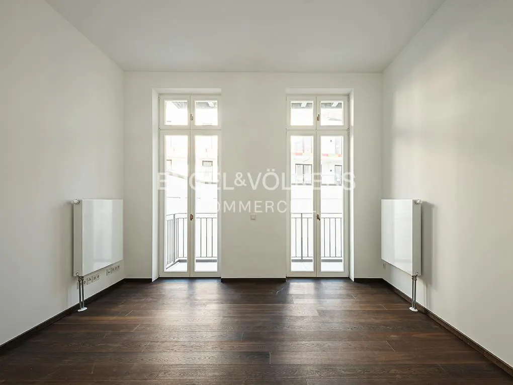 Empty room with dark wood floors, white walls, and two white doors leading to small balconies. White radiators are mounted on the walls.