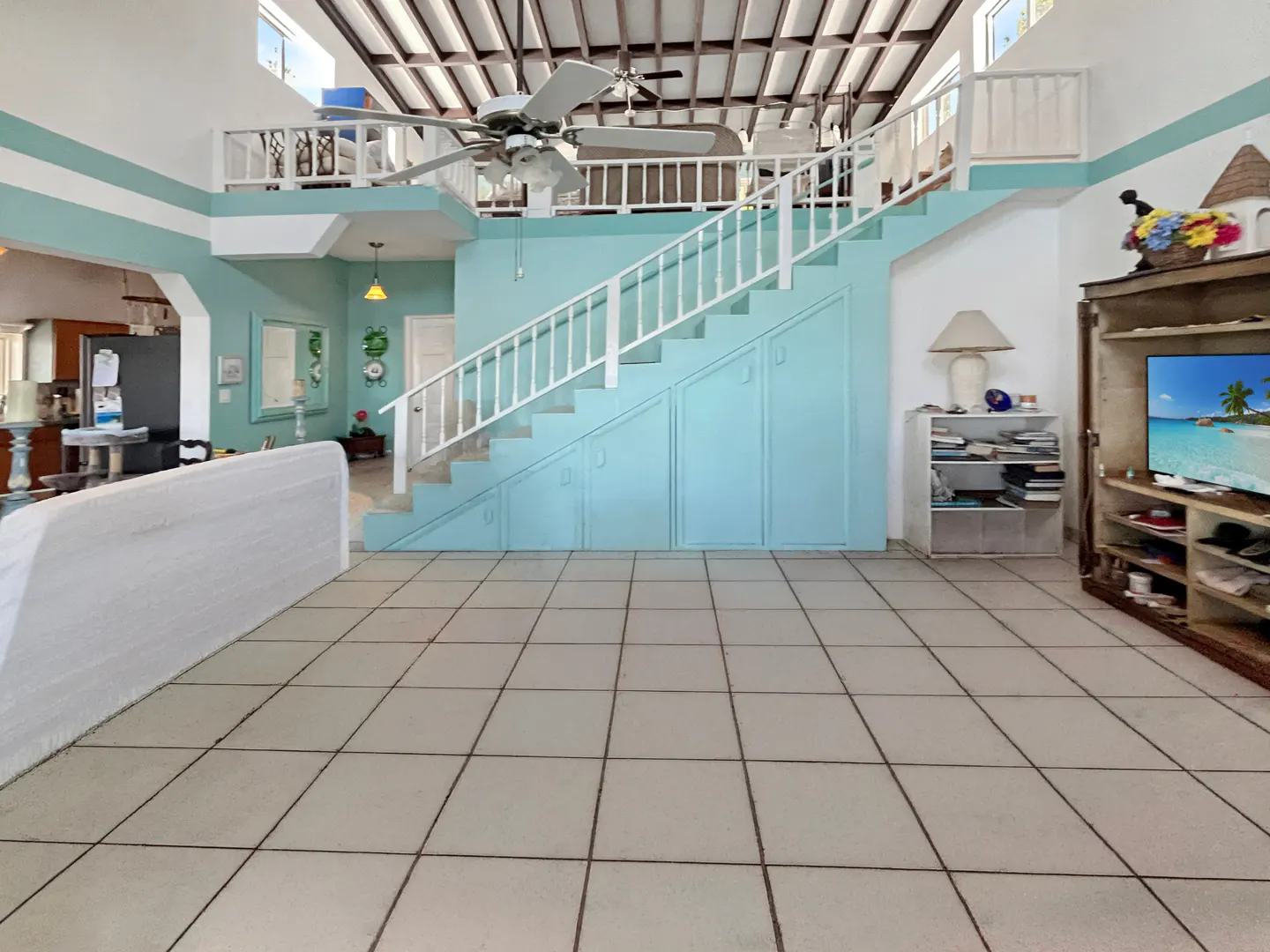 Bright, open living room with tile floors, blue walls, white stairs, and a loft. A TV and cabinet are on the right.
