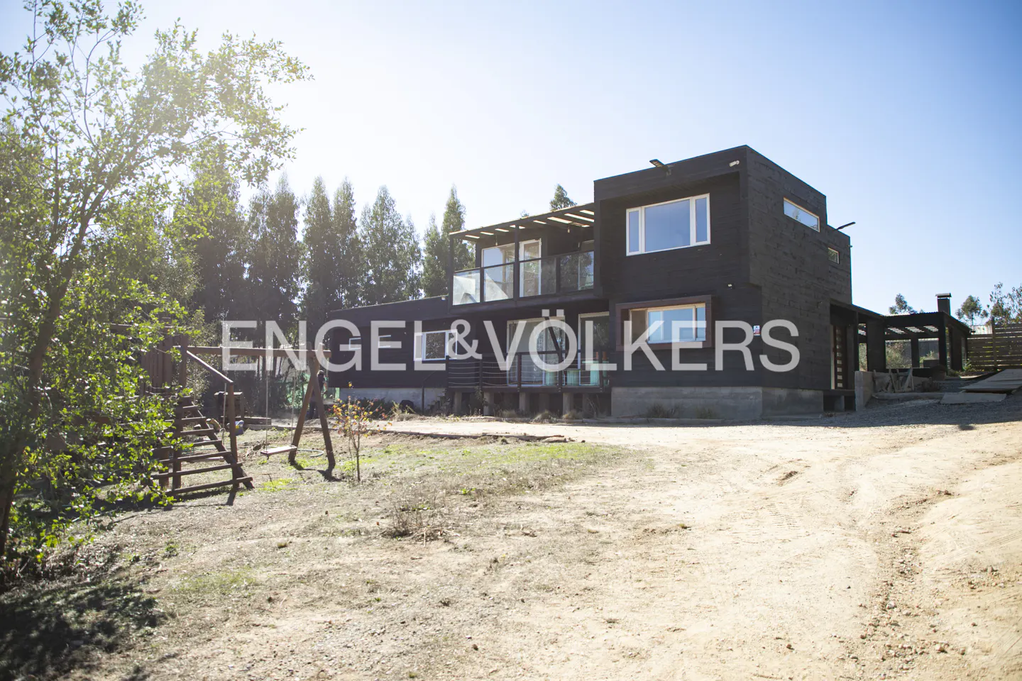 Two-story modern black house with balconies, surrounded by trees and a dirt yard under a clear blue sky. Engel & Völkers logo is superimposed.