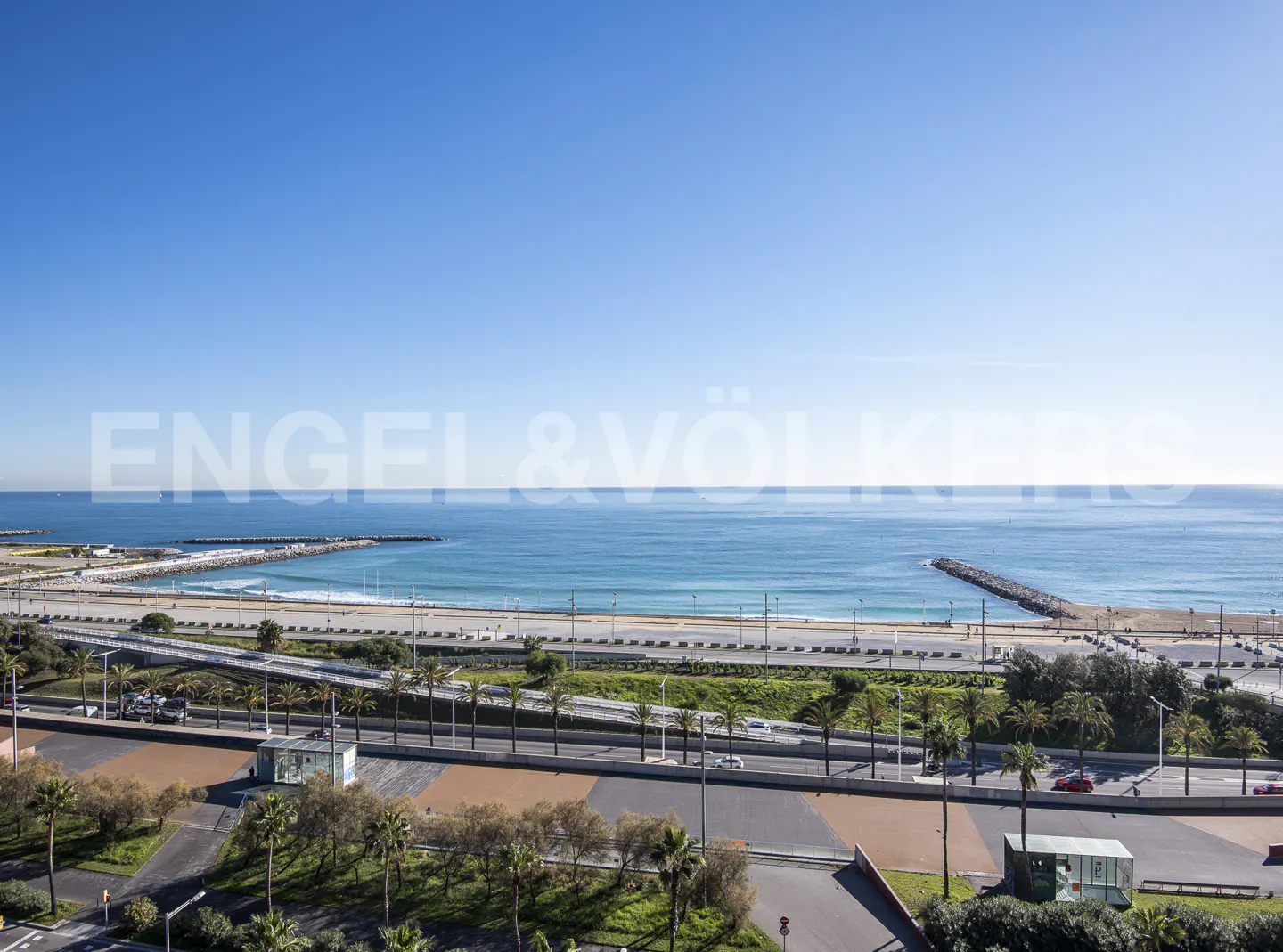 Ocean view with blue sky, beach, and palm trees. A road runs parallel to the beach. Engel & Völkers logo is superimposed.