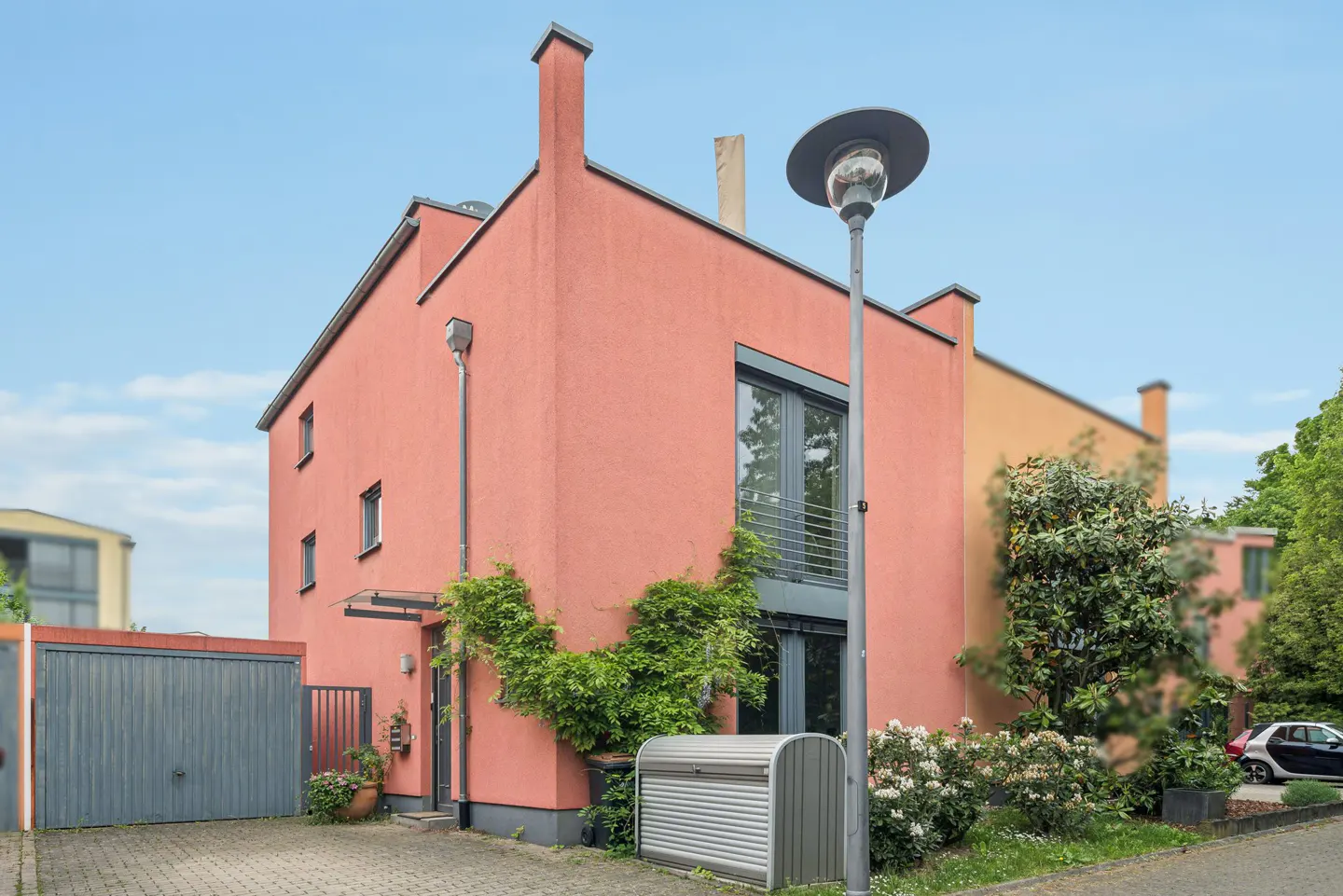 Modern salmon-colored house with a grey garage, climbing vines, and a street lamp against a clear blue sky.