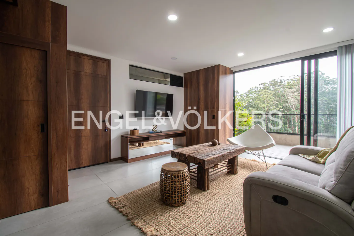 Living room with a gray sofa, wooden coffee table, and a white chair. Large windows overlook lush greenery. "Engel & Völkers" logo visible.