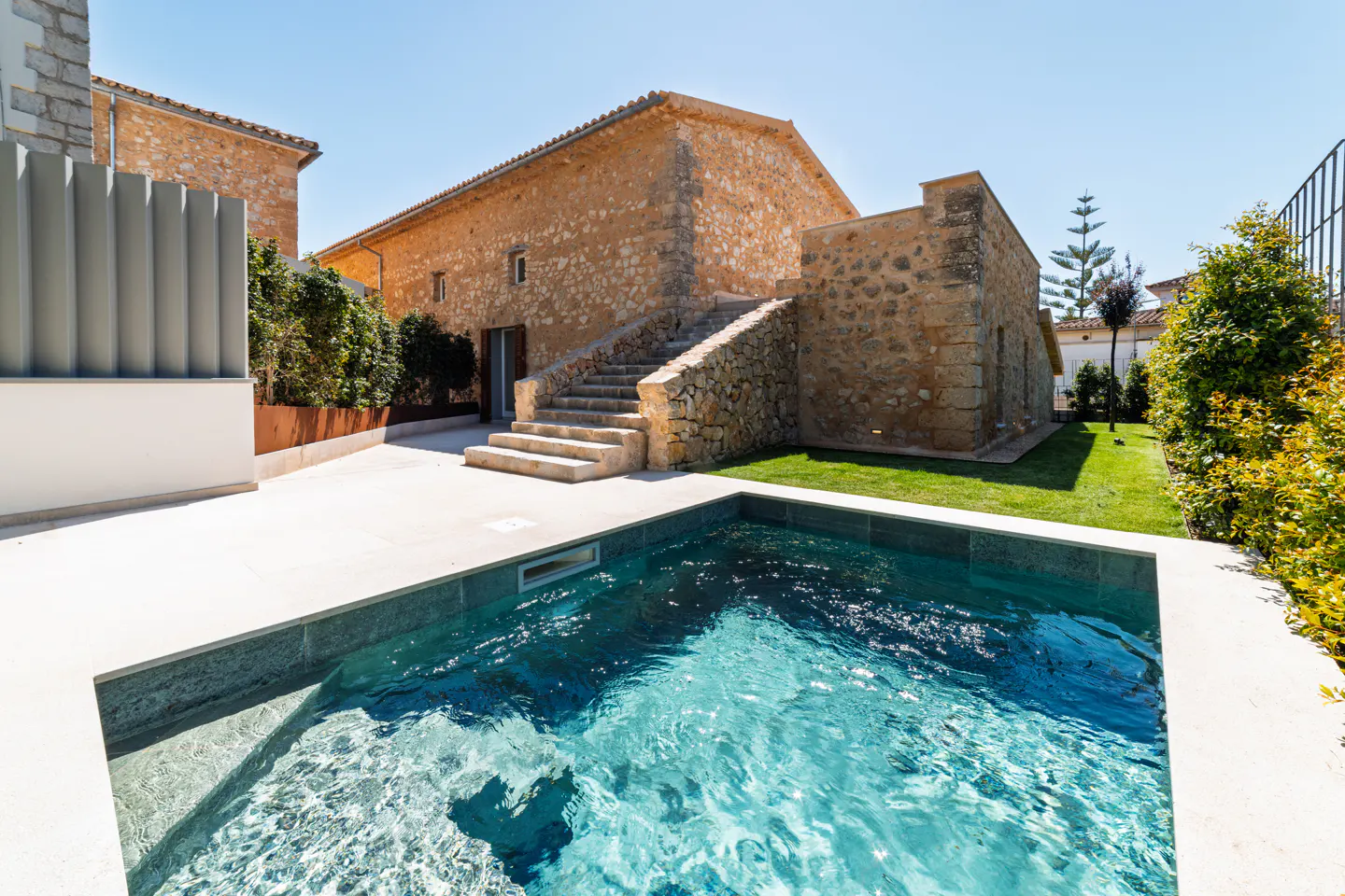 Exterior view of a stone house with a pool. The pool has clear blue water. Stone steps lead to the house. Green grass and plants surround the house.