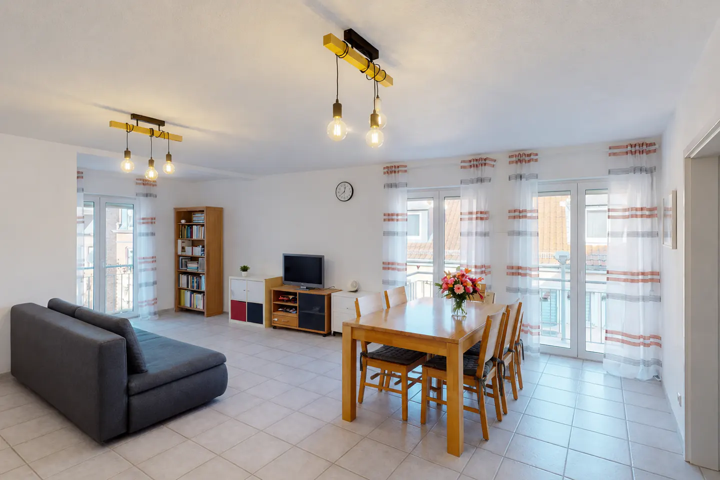 Bright living and dining area with gray sofa, wood table and chairs, TV, and bookshelf. Striped curtains adorn the windows.