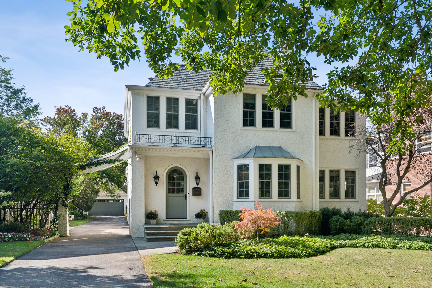 Exterior of a two-story white stucco house with a gray roof and a green front door. A driveway leads to a detached garage.