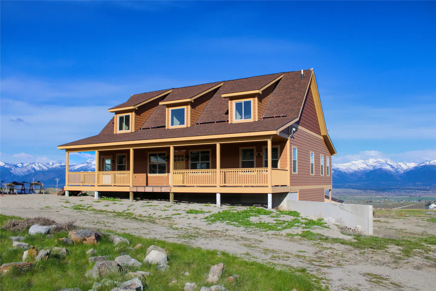 A two-story house with a porch, brown roof, and siding sits on a hill under a blue sky with mountains in the background.
