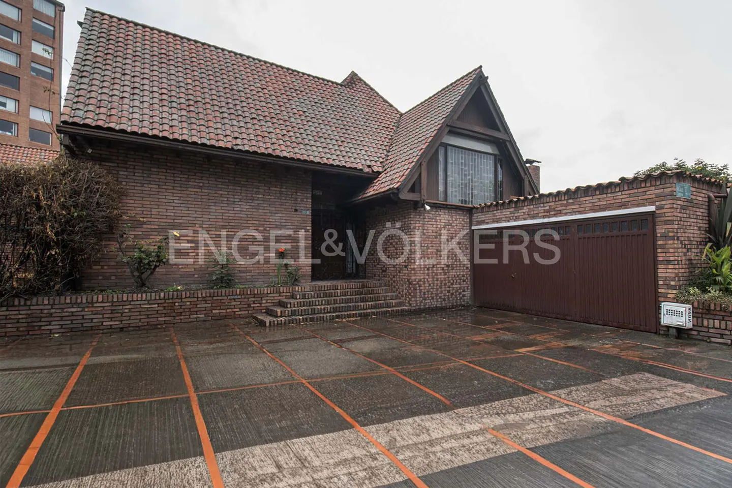 Exterior view of a brick house with a red tile roof and a brown garage door. The driveway is concrete with orange lines. Engel & Völkers logo is visible.