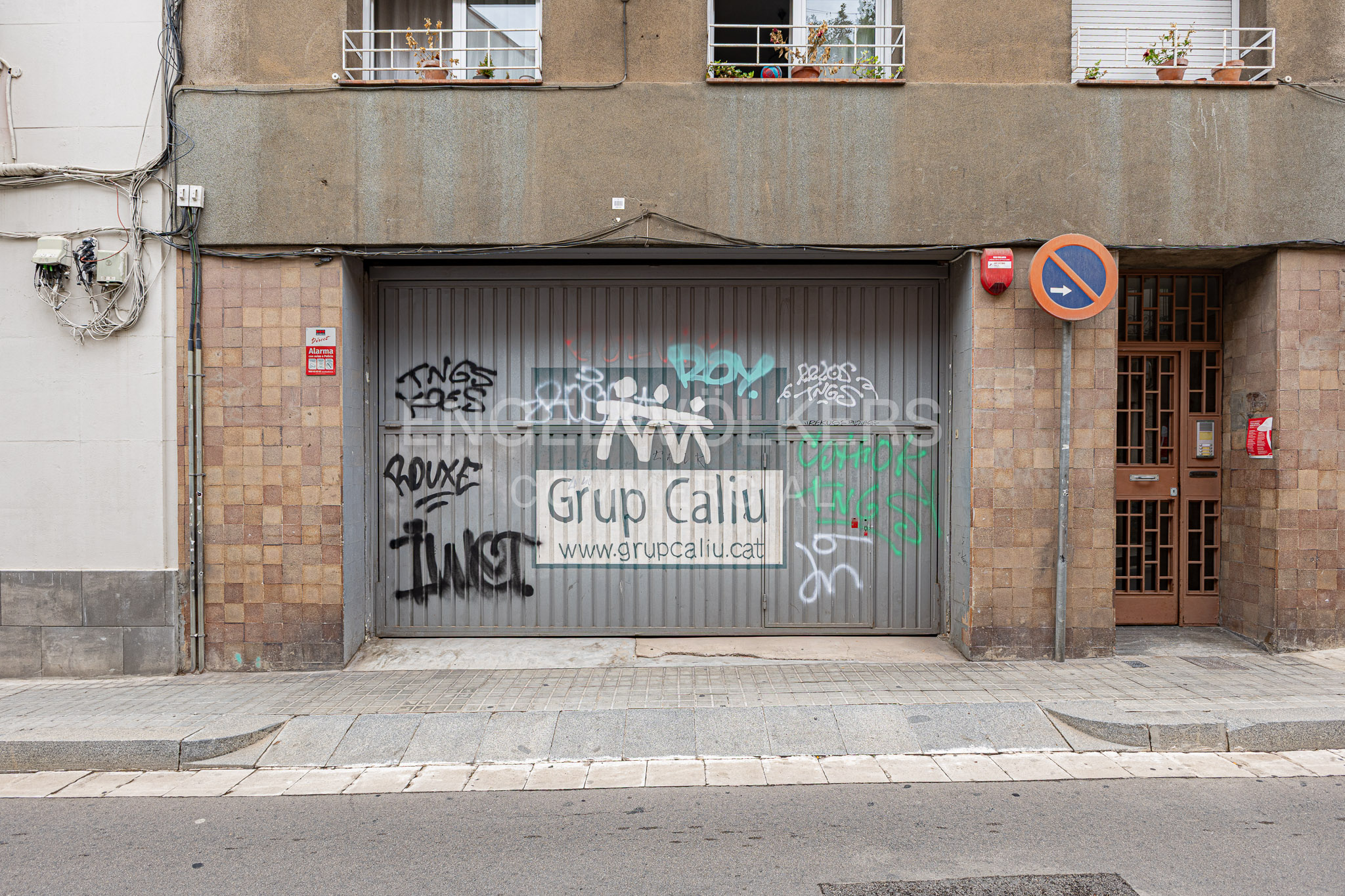 A gray metal garage door with graffiti and a sign for Grup Caliu, set in a brown tiled building facade on a city street. [AI-generated text]