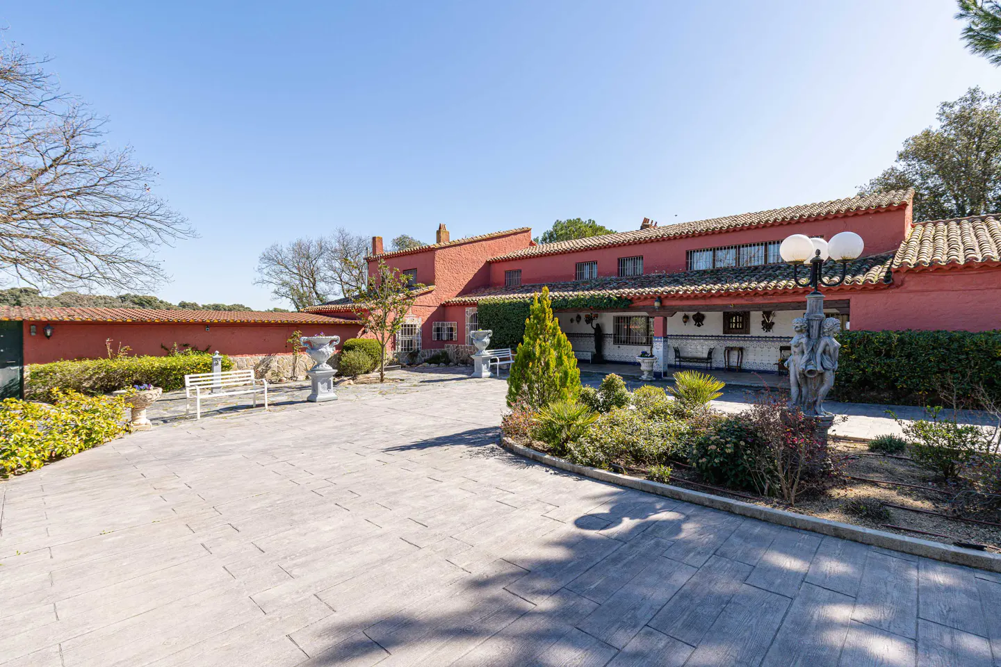 Exterior view of a red, single-story house with a tile roof, a stone courtyard, and manicured landscaping.