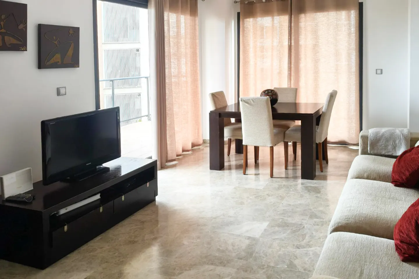 Living room with a dark wood TV stand, a dining table with four chairs, and a beige sofa with red pillows. The floor is marble.
