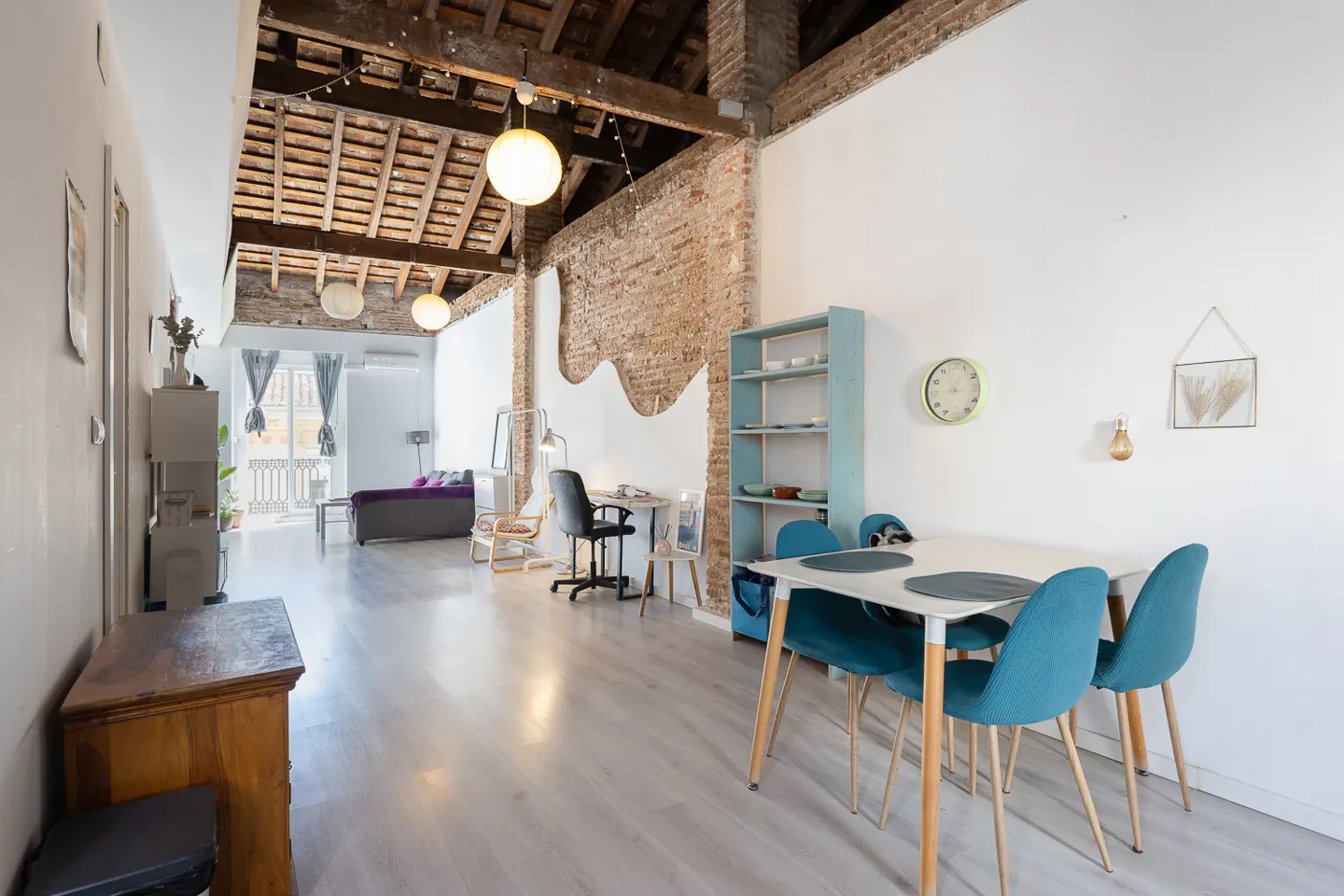 Bright, open loft apartment with exposed brick, wood beams, and white walls. A dining table with blue chairs sits near a desk and bookshelf.