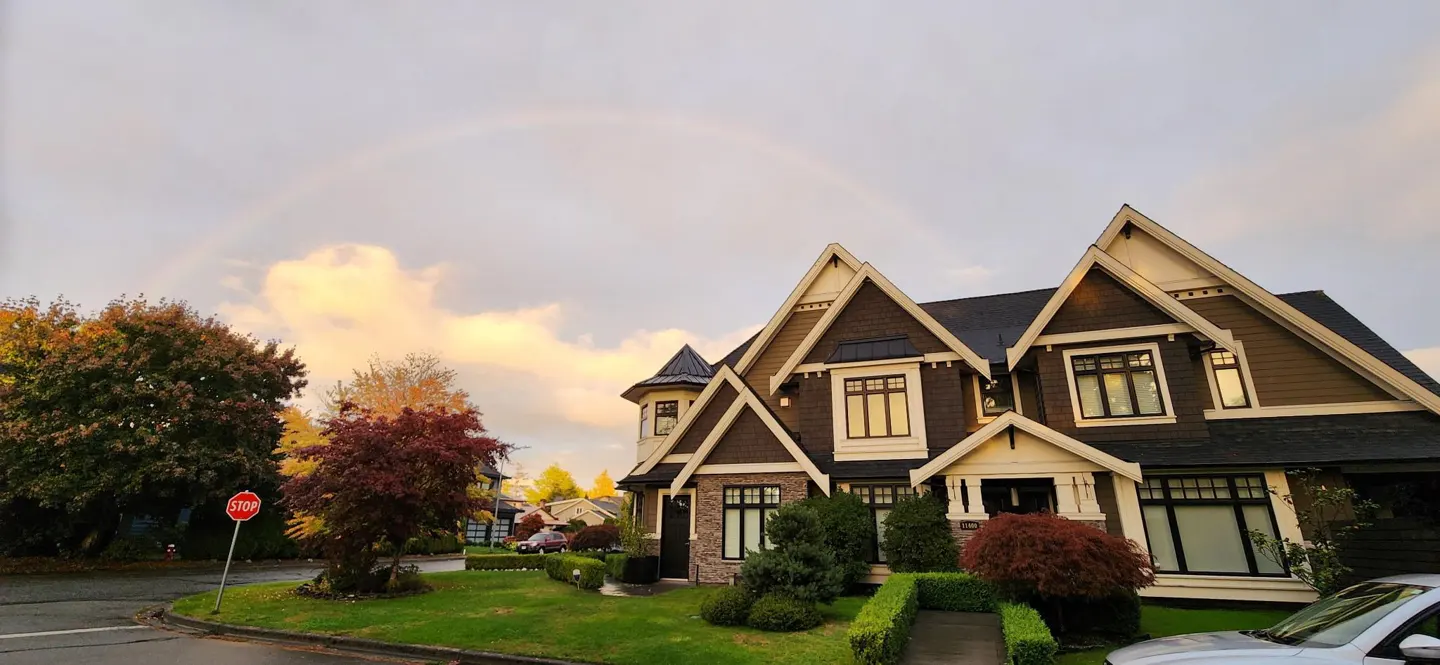 A brown house with white trim sits under a rainbow, with green grass and trees in autumn colors.