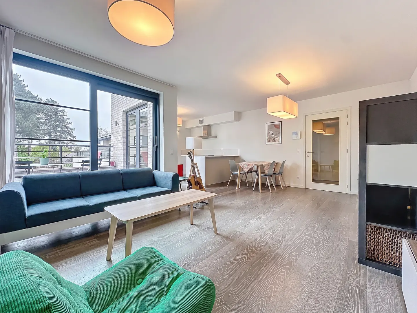 A bright, open-concept living space with a blue sofa, wood floors, and a view of a balcony. A guitar leans against a kitchen island.