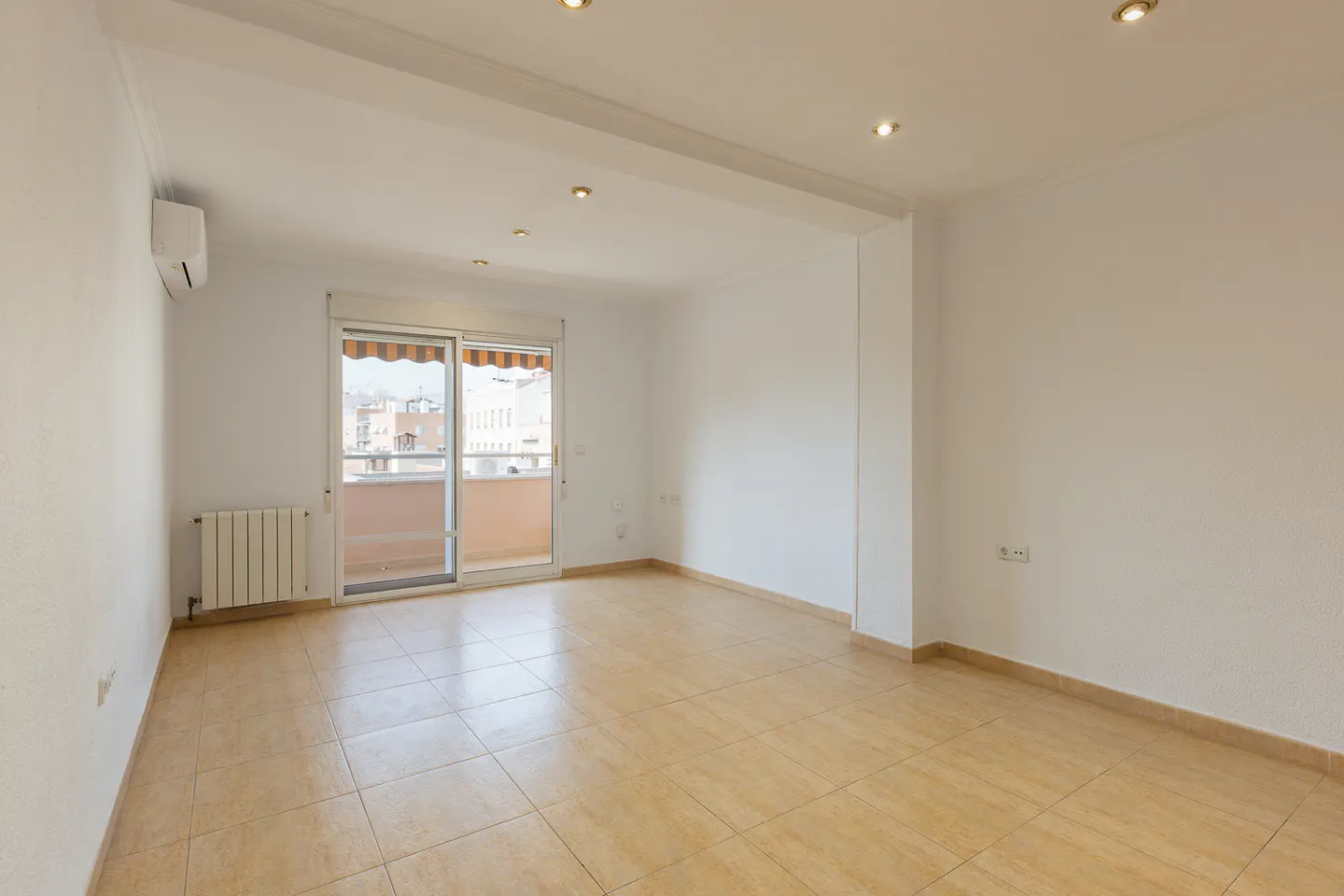 Bright, empty room with beige tile floor, white walls, and recessed lighting. Sliding glass doors lead to a balcony with a striped awning.