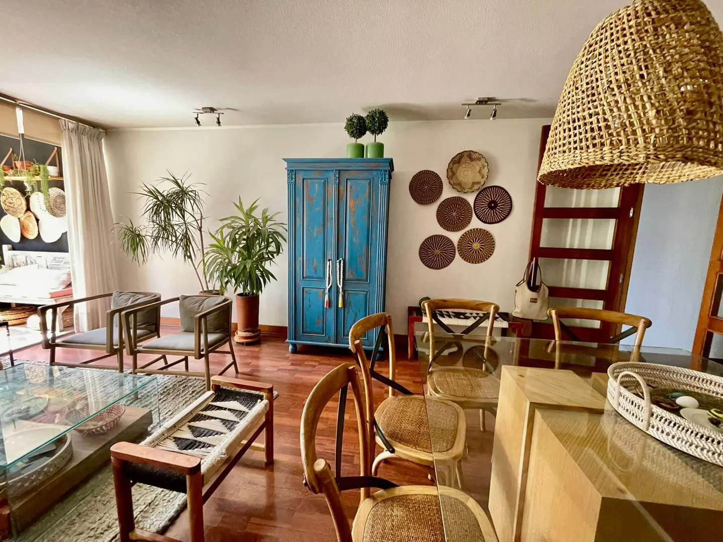 A bright living room with wood floors, chairs, plants, and a blue cabinet. Woven wall art and a wicker light fixture add texture.