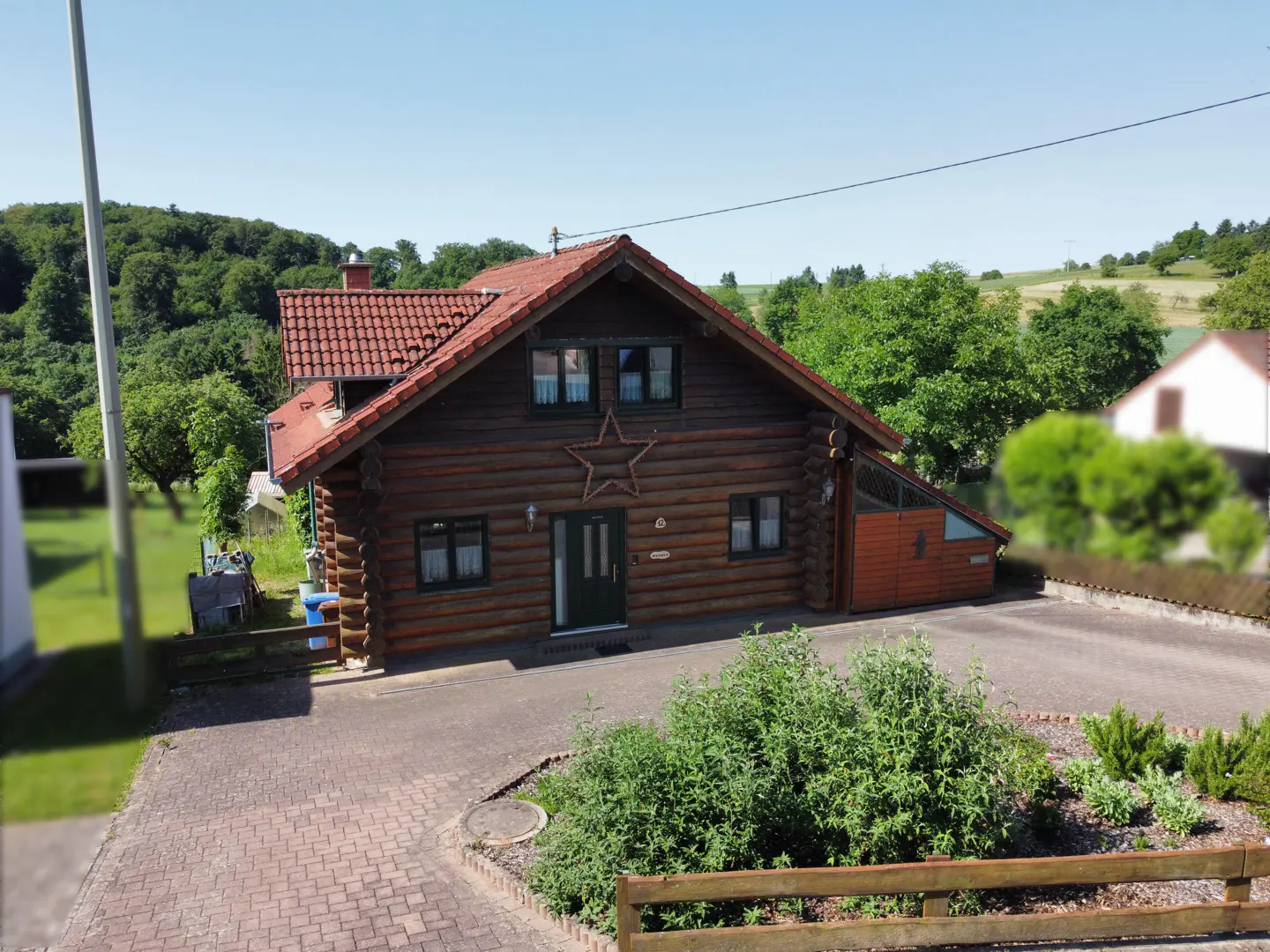 A rustic brown log cabin with a red tiled roof, a star decoration on the front, and a paved driveway in a rural setting.