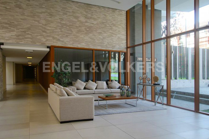 A modern lobby with a beige sofa, coffee table, and floor-to-ceiling windows. The wall is textured beige brick. Engel & Volkers logo is visible.