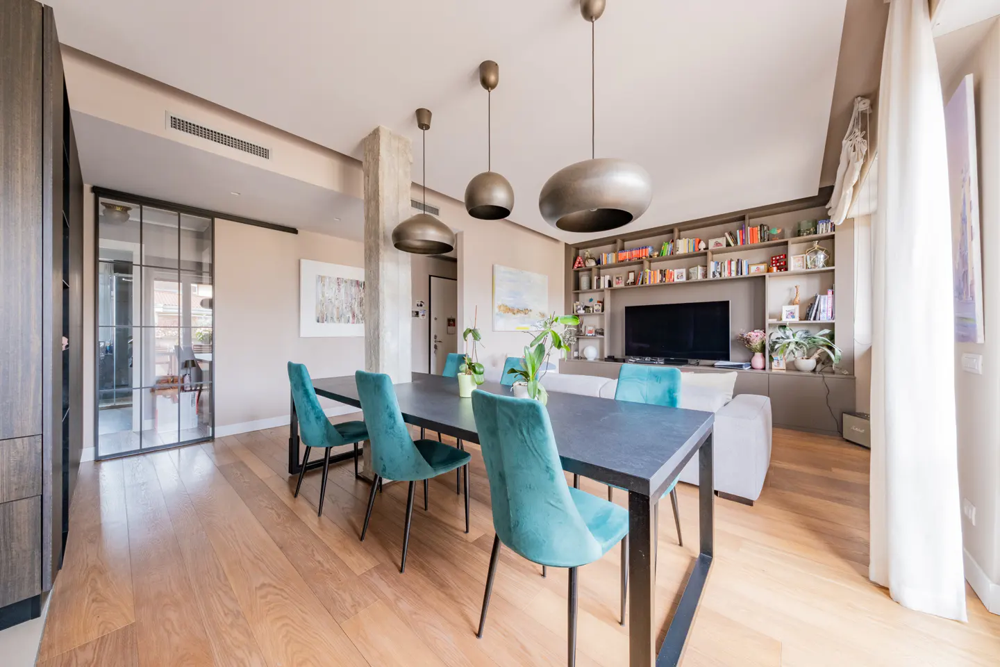 Modern dining area with a black table, teal velvet chairs, pendant lights, and a living room with a TV and bookshelf.