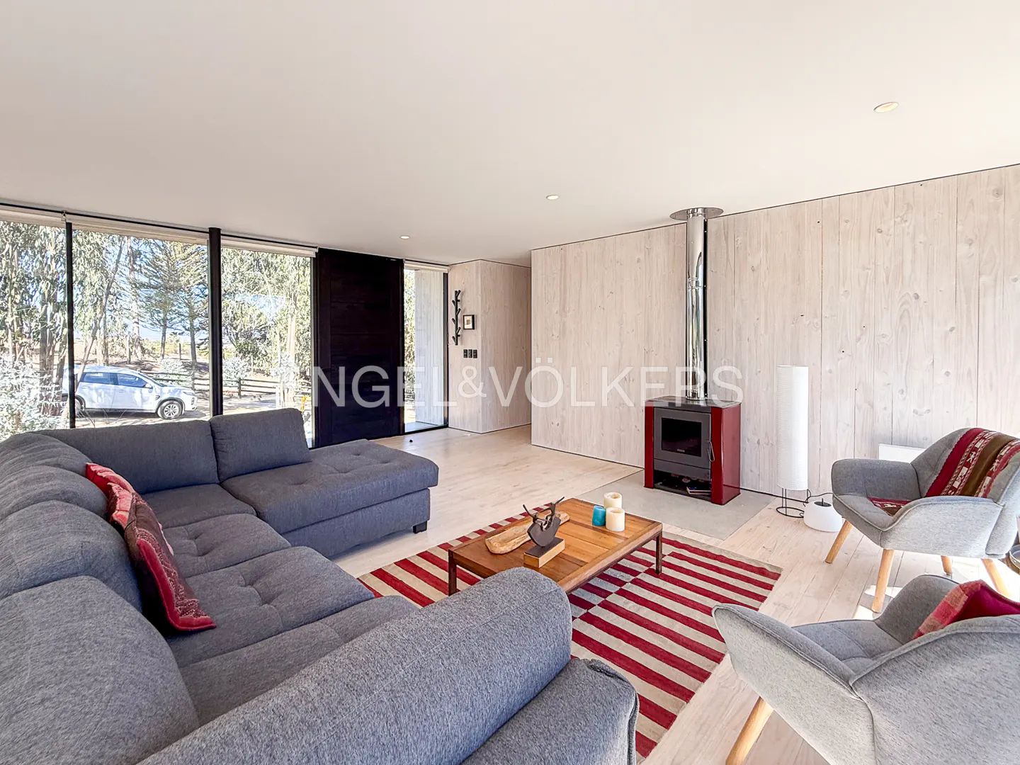 Bright living room with gray sectional sofa, wood coffee table on red/white striped rug, and red wood-burning stove with metal chimney.