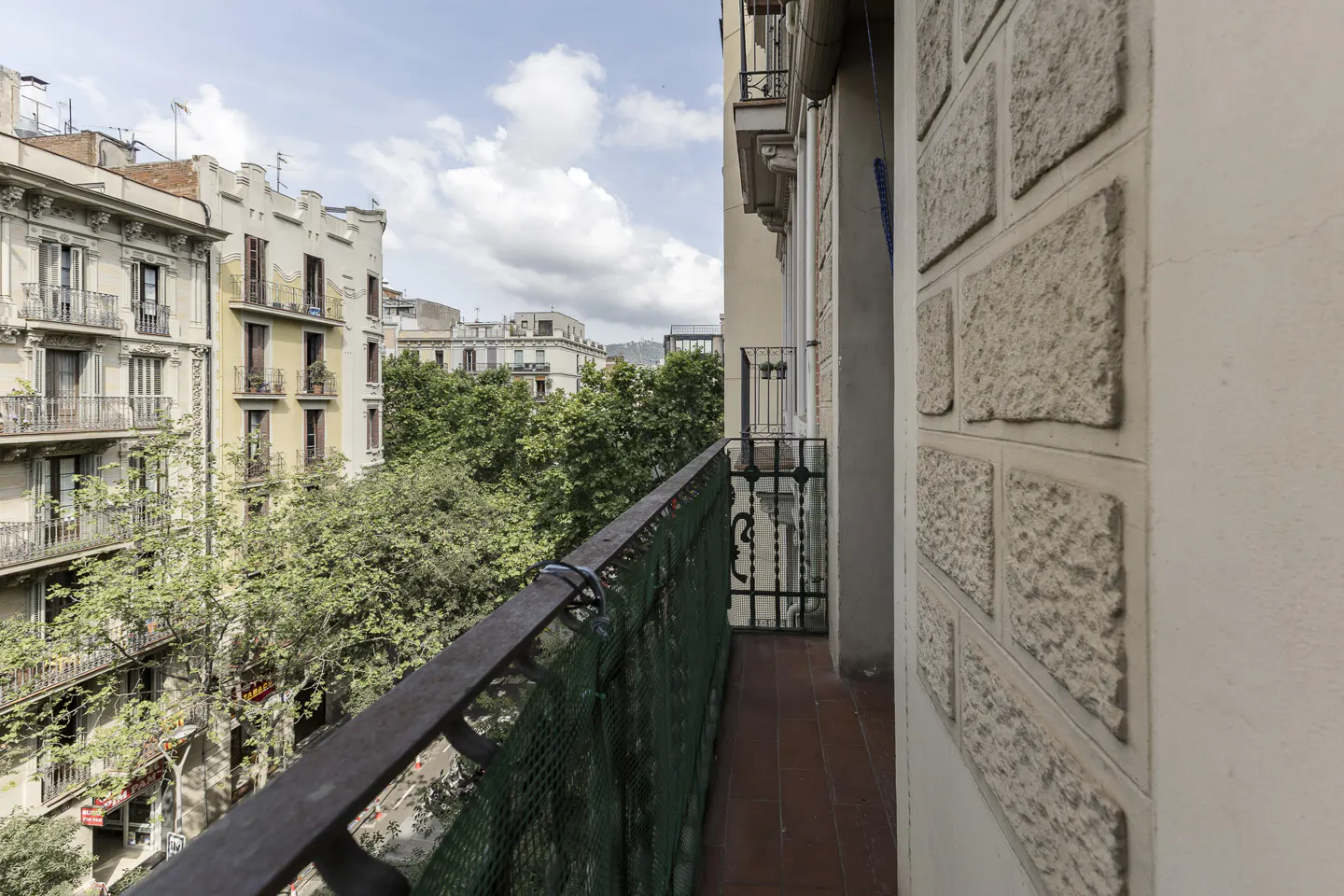 View from a balcony with a dark metal railing and green mesh, overlooking buildings and trees in a city.