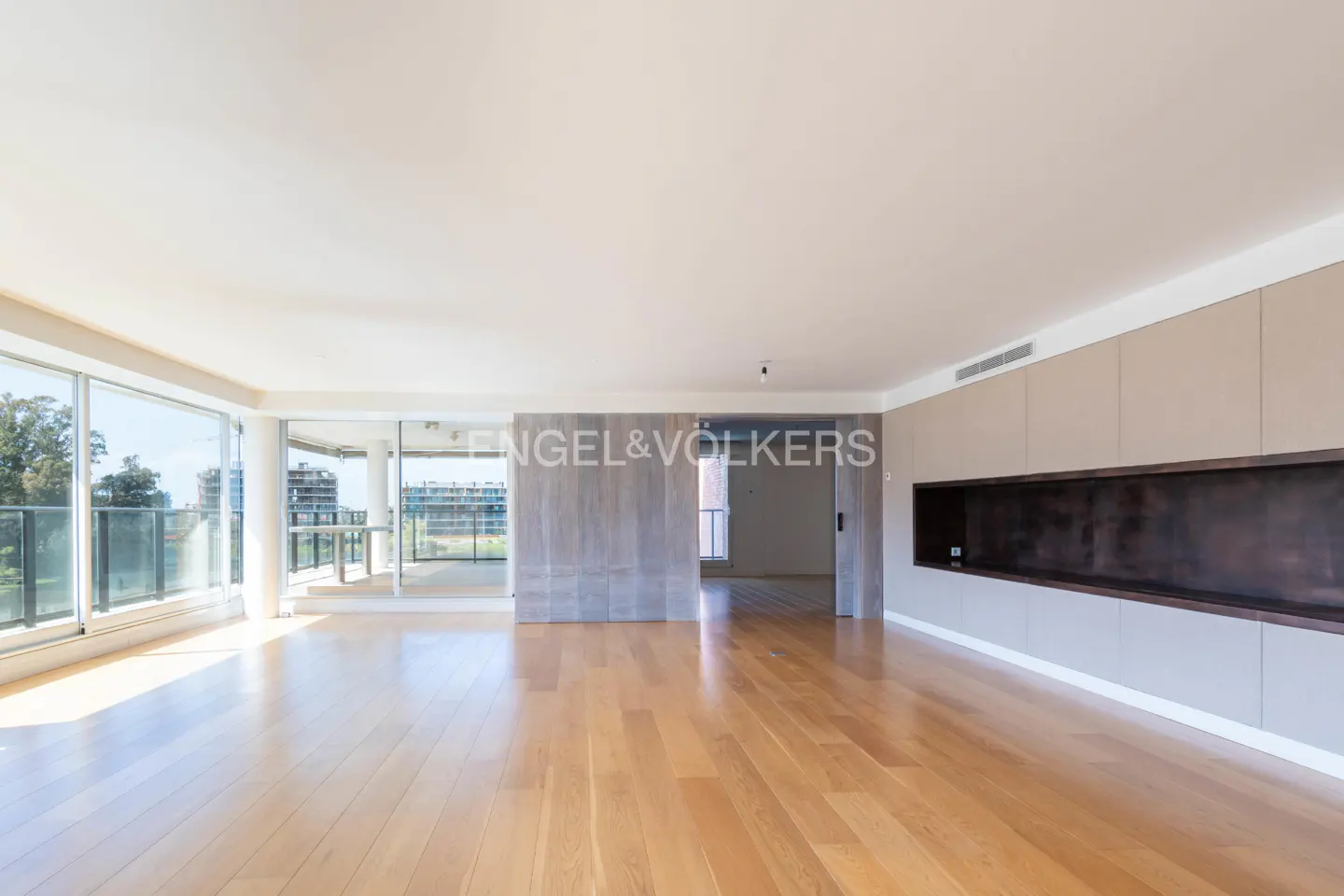 Bright, empty room with wood floors, large windows, and a balcony. Built-in cabinets and a dark recessed shelf line one wall. Engel & Völkers logo.