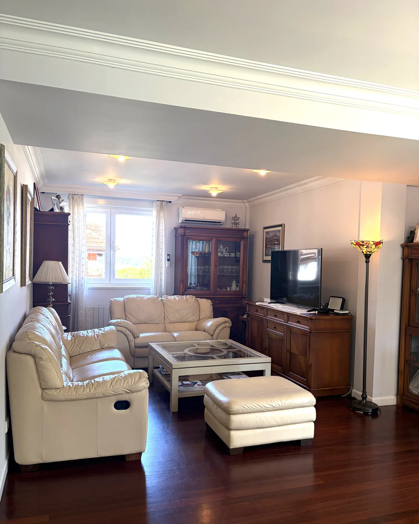 Living room with cream leather sofas, wood floors, and a large window. A glass-topped coffee table sits in the center.