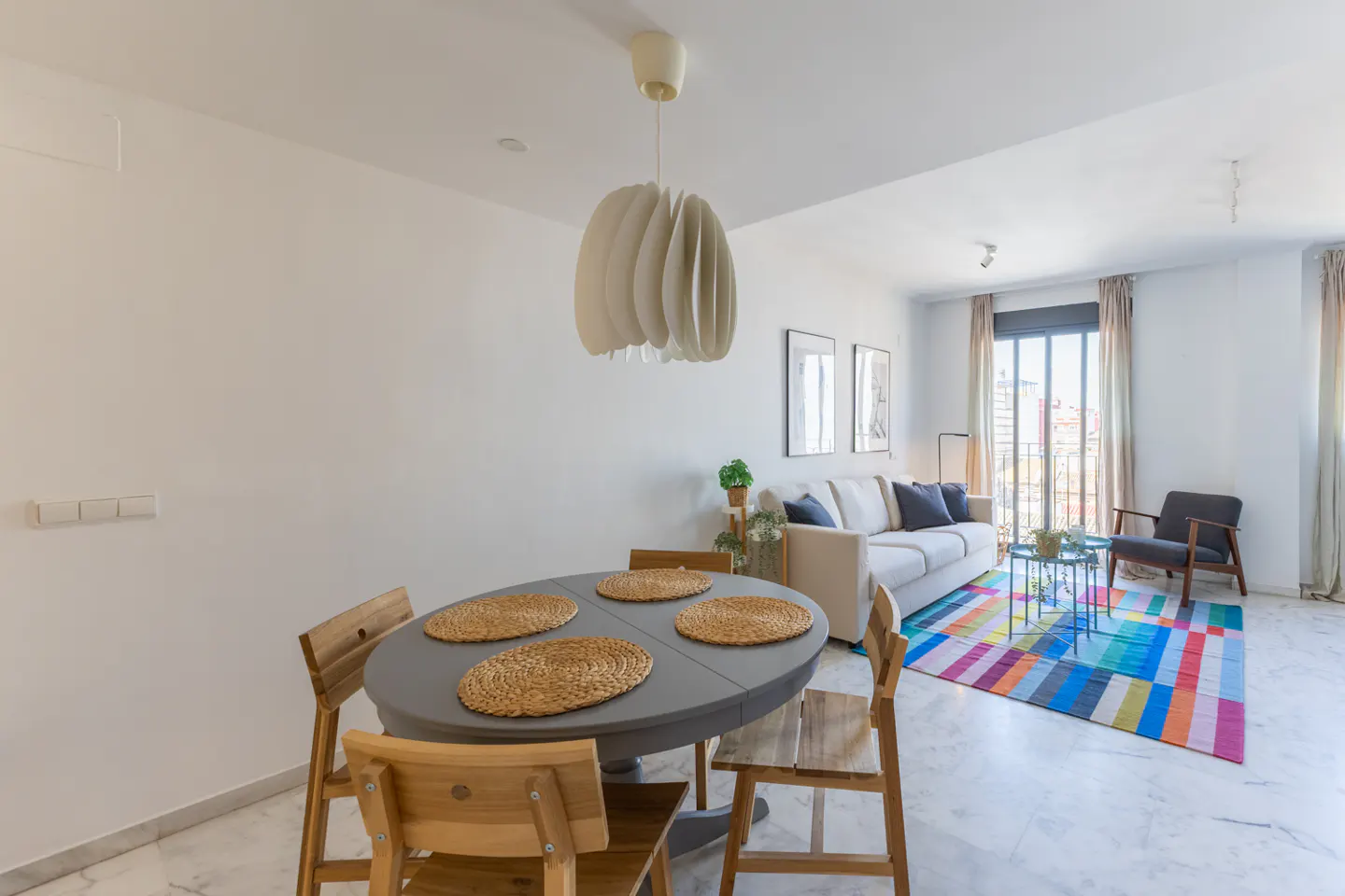 Bright living room with a gray oval table, wooden chairs, and a colorful striped rug. A white sofa sits near a window.