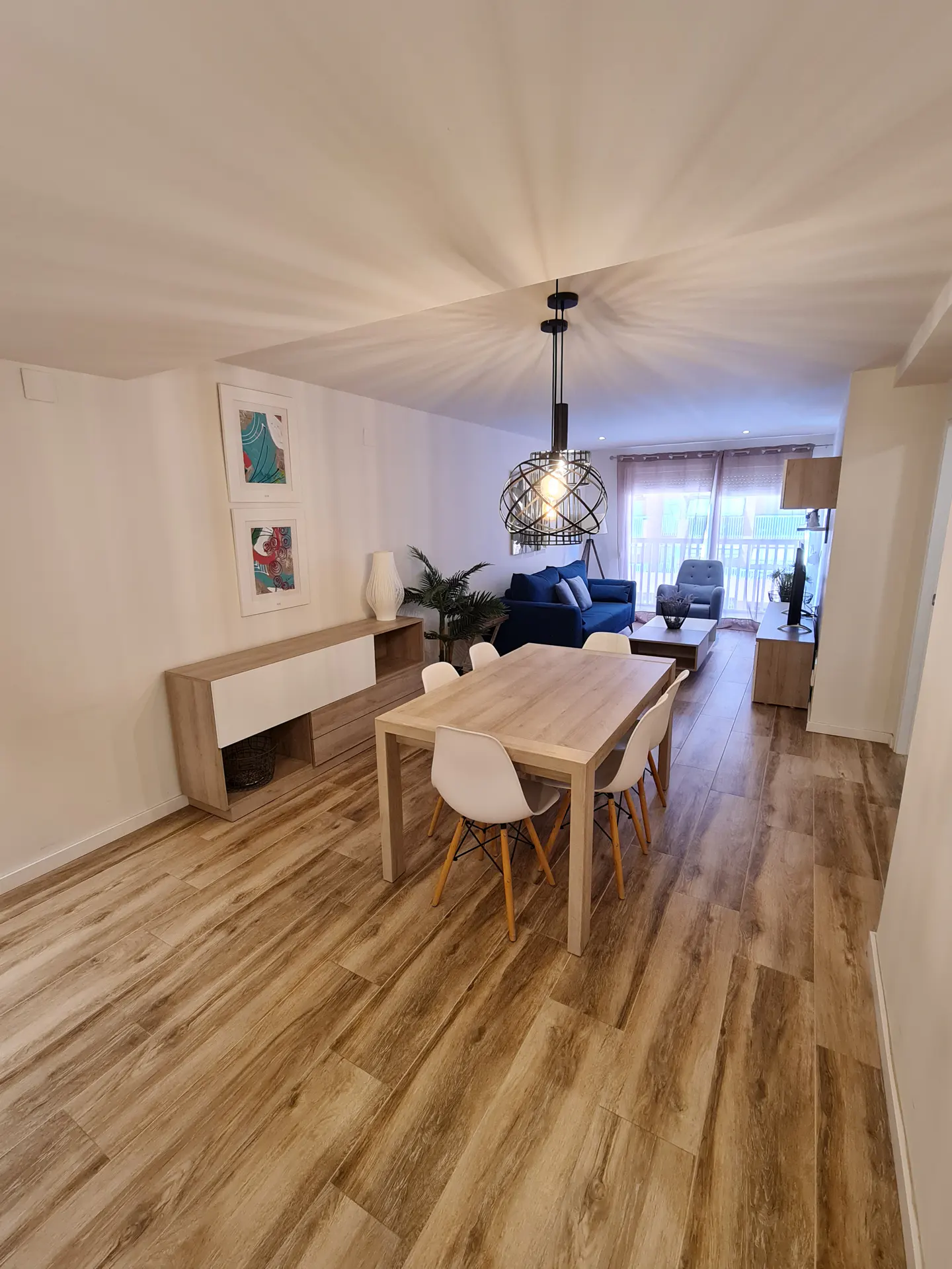 Open-concept living and dining area with wood-look tile flooring. A light wood table with white chairs sits under a modern chandelier. Blue sofa in the background.