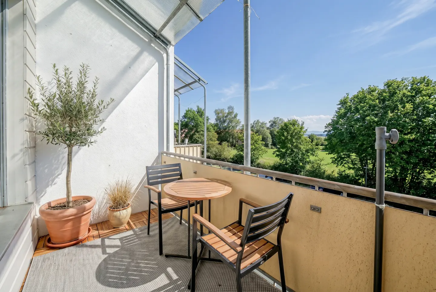 Balcony with a round wooden table and two chairs, potted plants, and a view of green trees and blue sky.