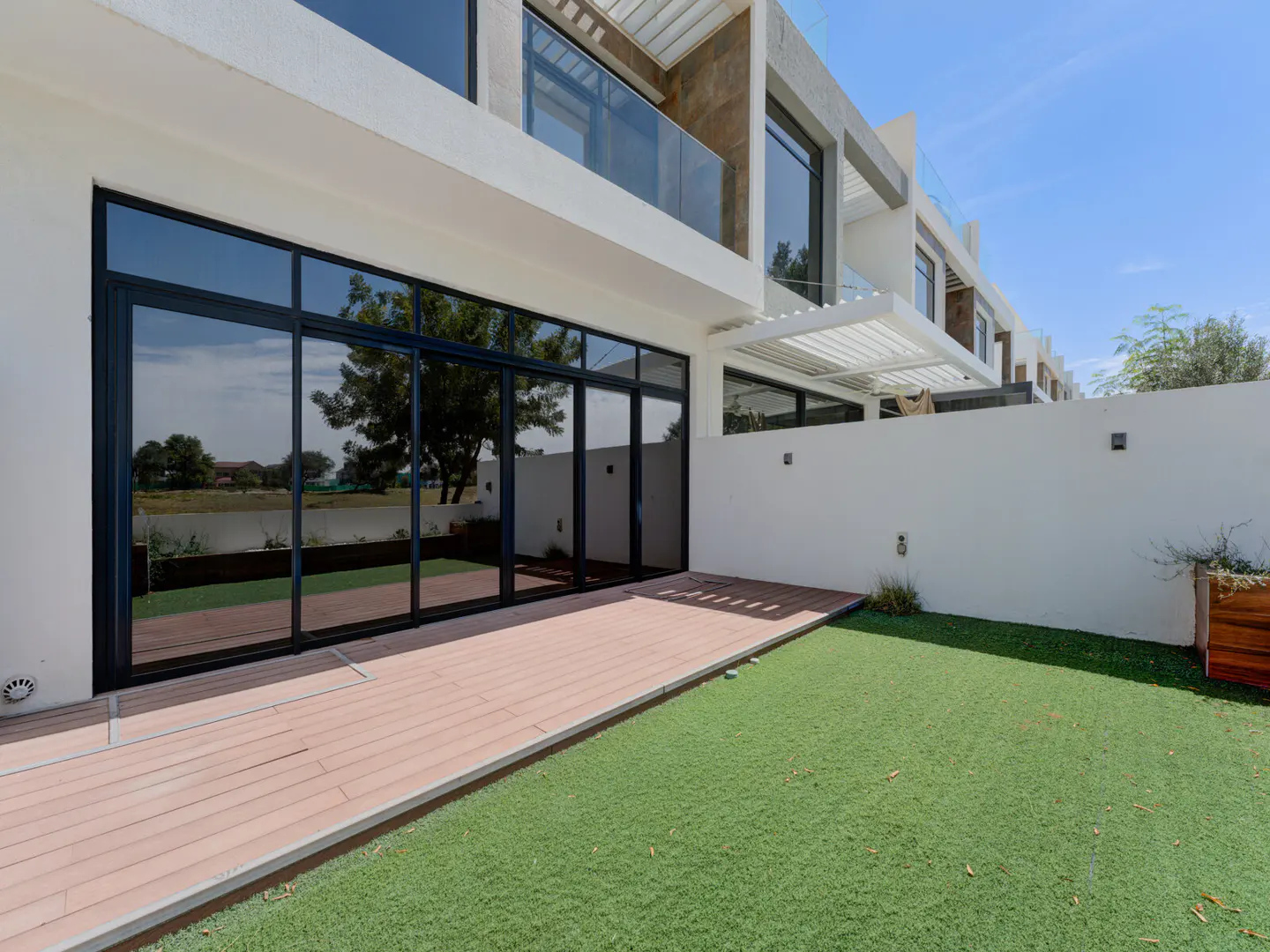Modern house exterior with a green lawn, wooden patio, and large black-framed sliding glass doors reflecting trees.