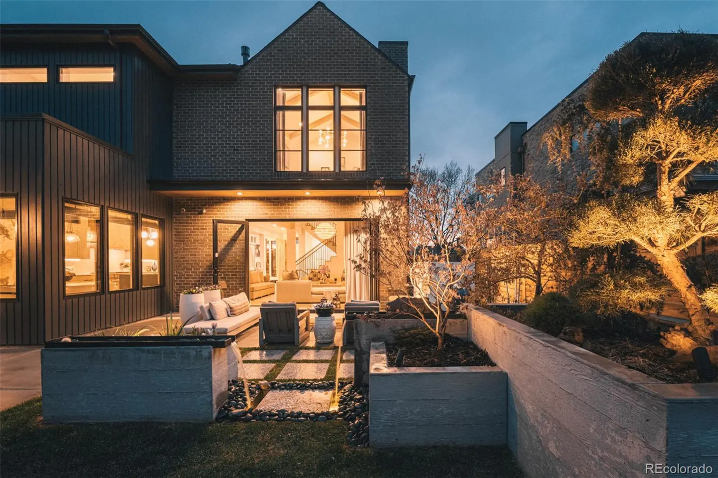 Backyard view of a modern brick home with a patio, furniture, and illuminated trees at dusk. Interior visible through open doors.