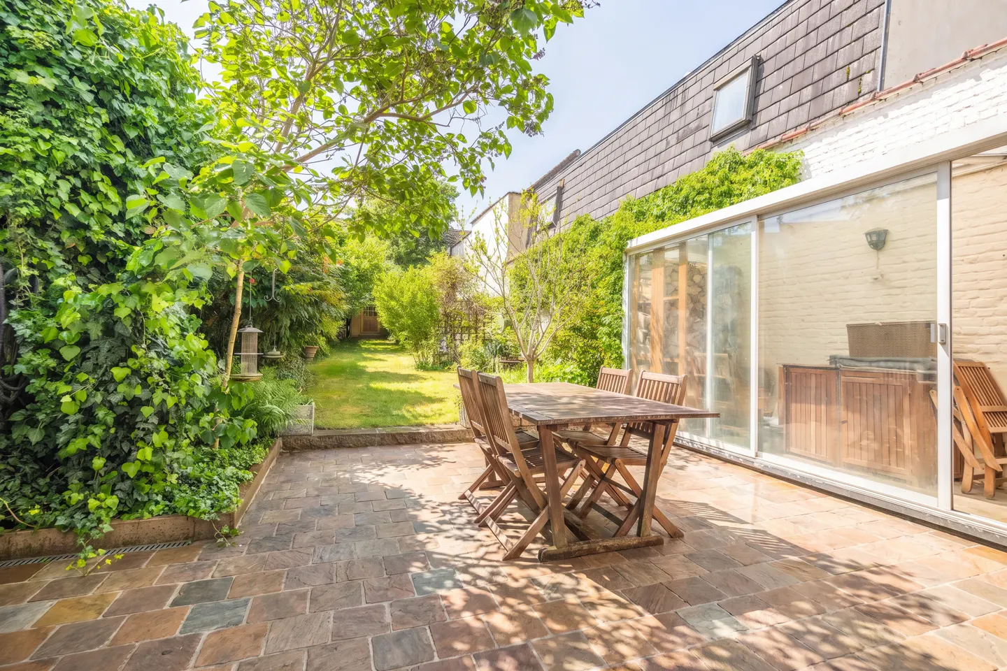 A backyard patio with a wooden table and chairs, a green lawn, and a building with a glass-enclosed porch.