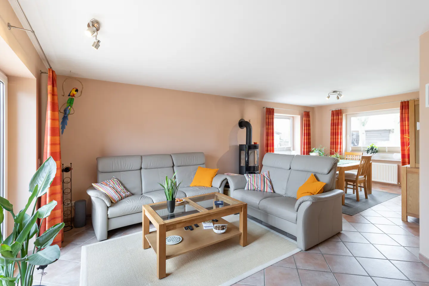 Living room with gray sofas, wood table, and black fireplace. Orange curtains frame windows. Dining table visible in background.