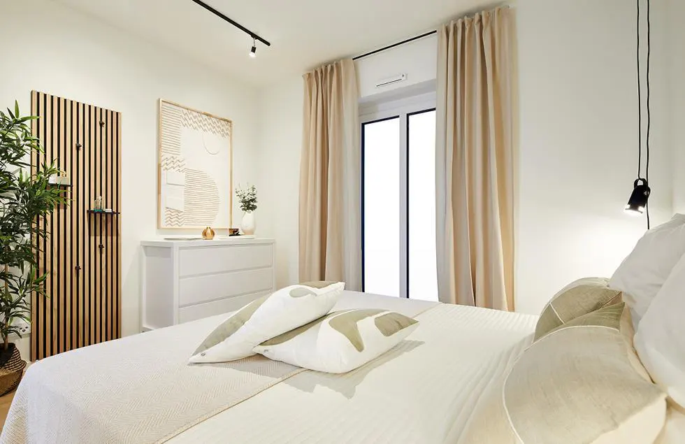 A bright bedroom with a white bed, beige curtains, and a white dresser. A wooden slat wall and a plant add texture. A modern art piece hangs above the dresser.