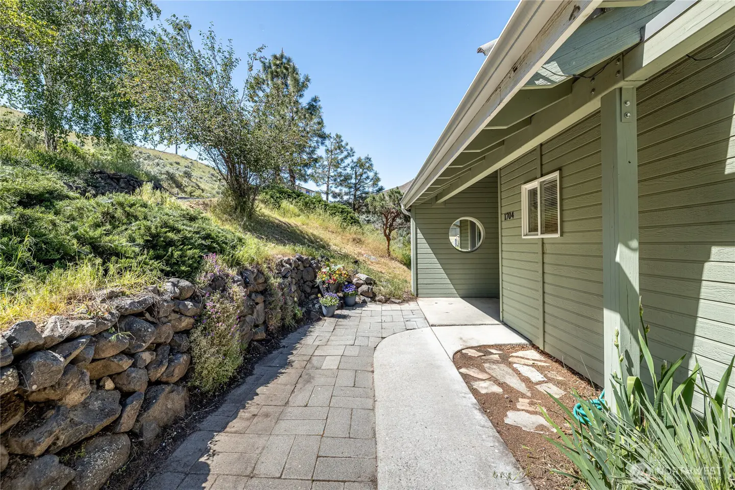 Exterior of a green house with a stone walkway and hillside landscaping. A round window is visible.