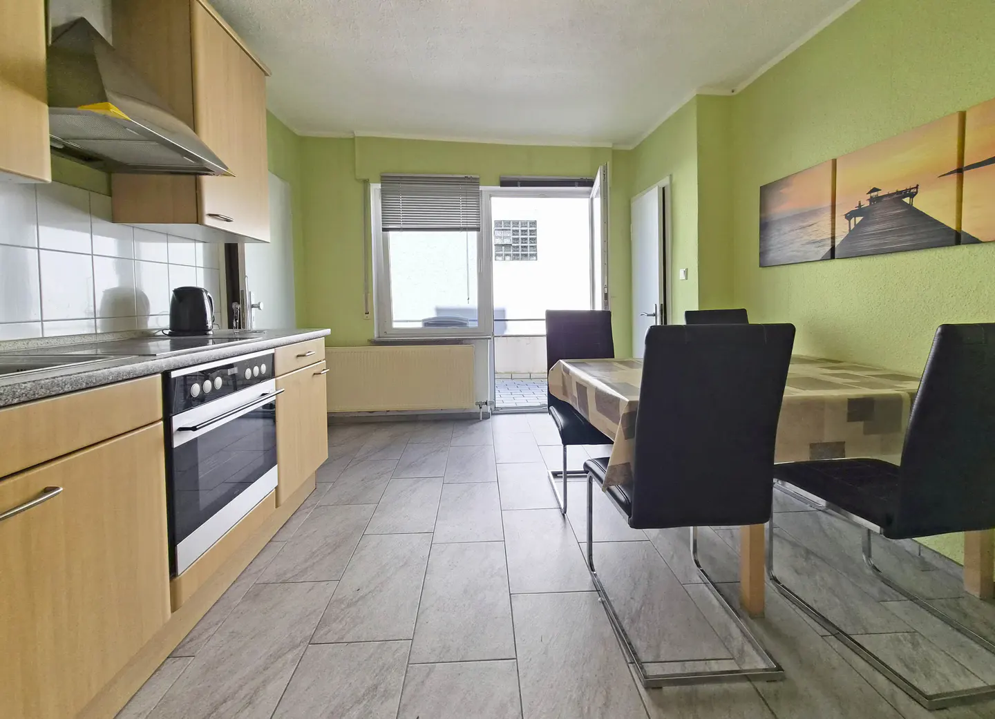 A kitchen and dining area with light green walls, light wood cabinets, and a table with black chairs. A balcony is visible through an open door.