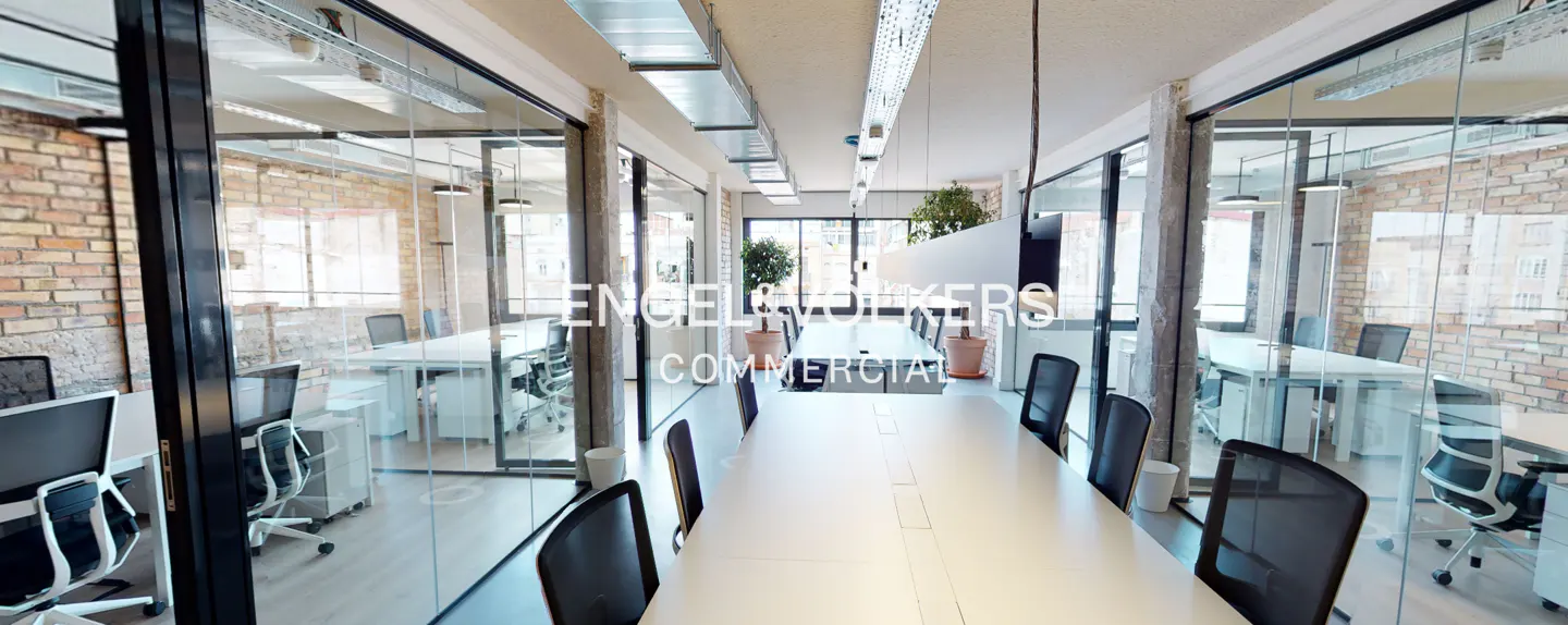 Bright office space with a long white table and black chairs. Glass-walled offices with brick walls are on either side. "Engel & Volkers Commercial" is on the glass.