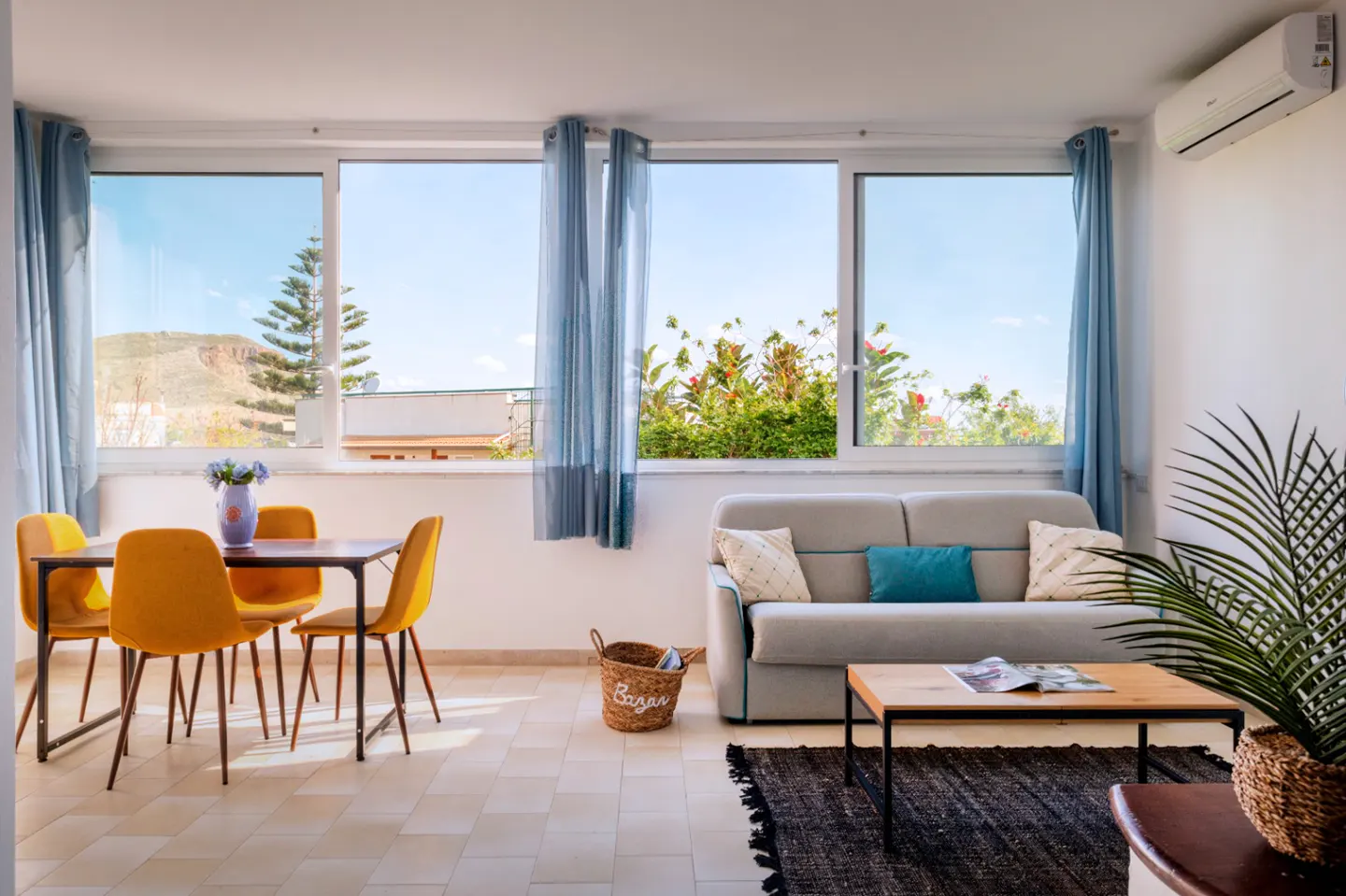Bright living room with a dining table, yellow chairs, a gray sofa, and a view of greenery and blue sky through large windows.