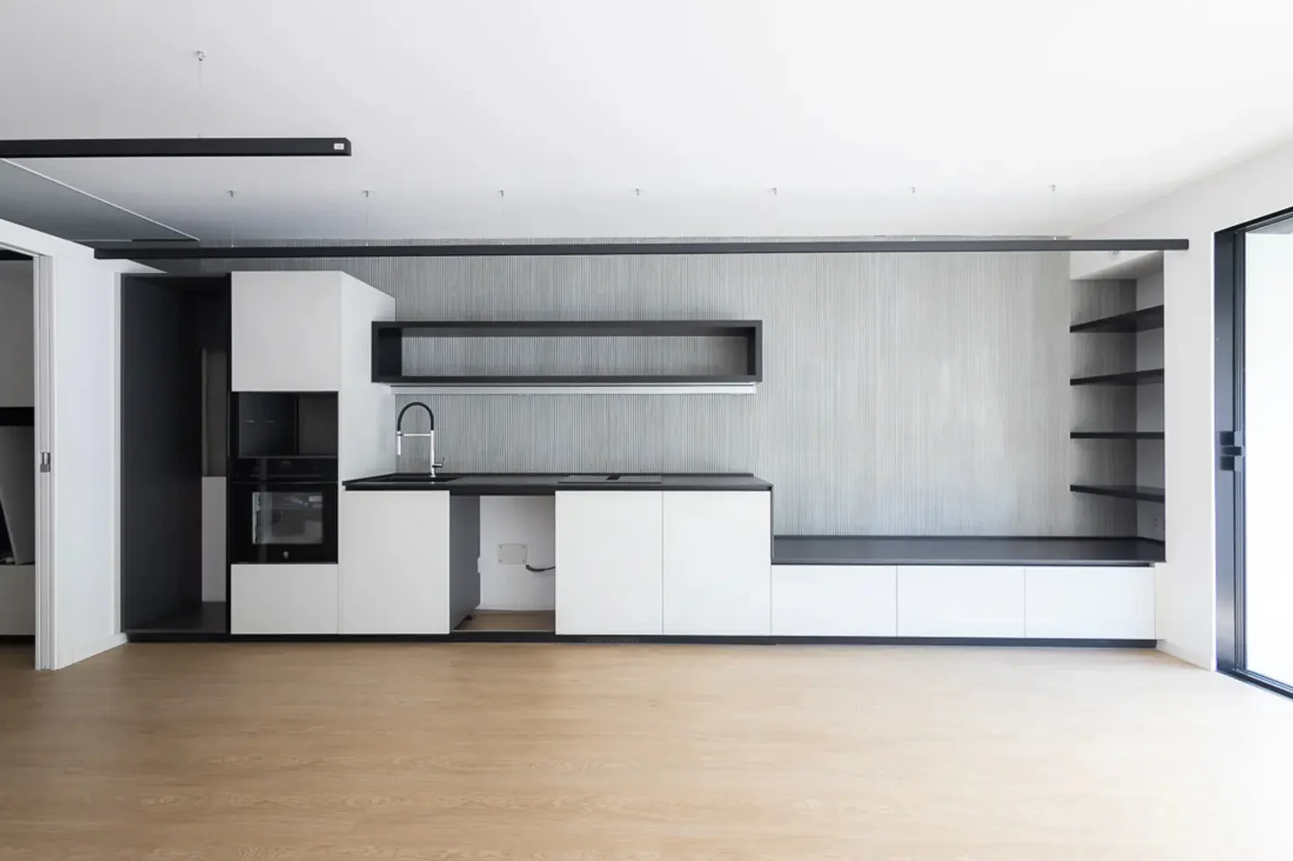 Modern minimalist kitchen with white cabinets, black accents, grey textured wall, and light wood flooring.