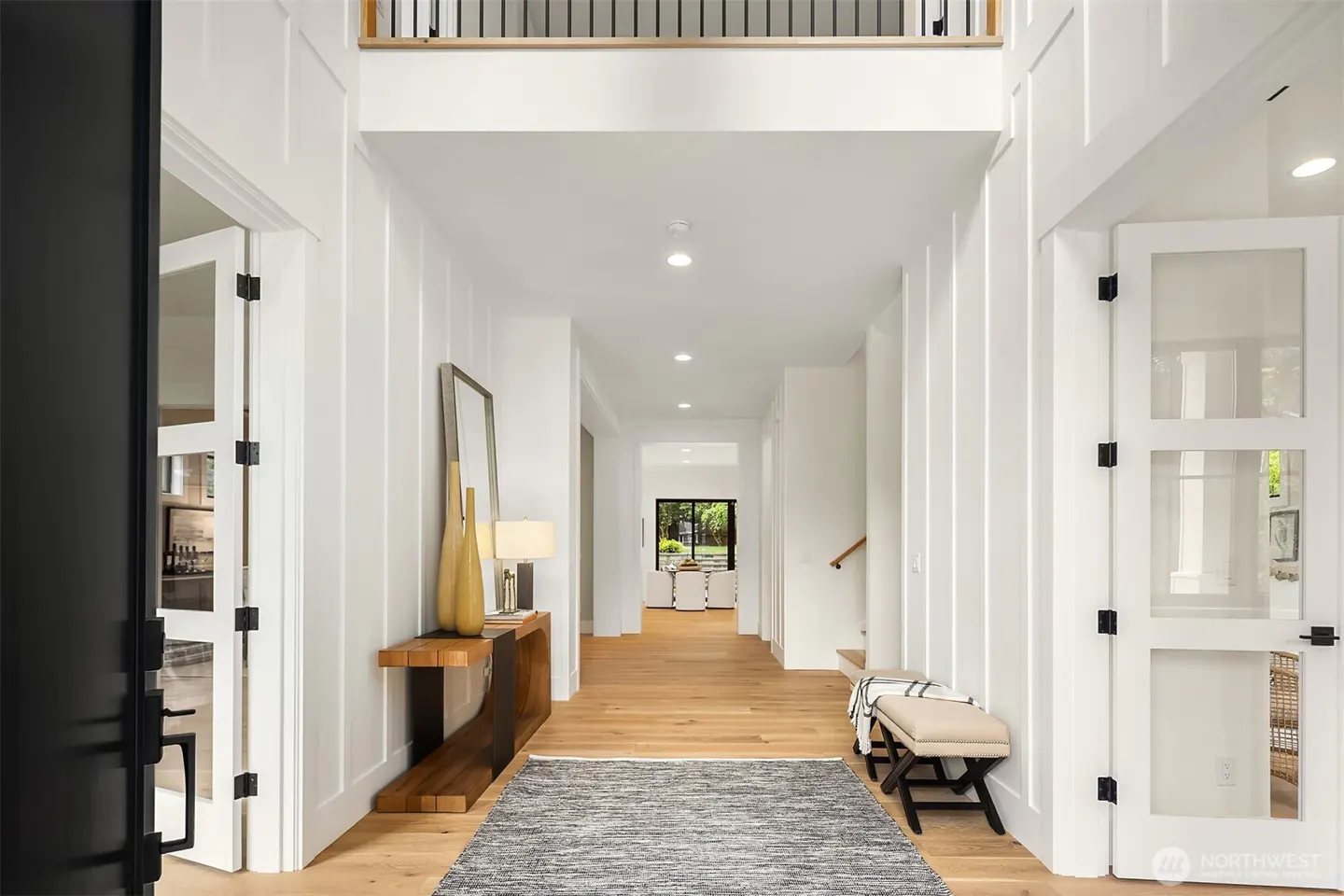 Bright foyer with white walls, wood floors, and a gray rug. A wooden console table with lamps and decor sits to the left. Glass-paneled doors add light.