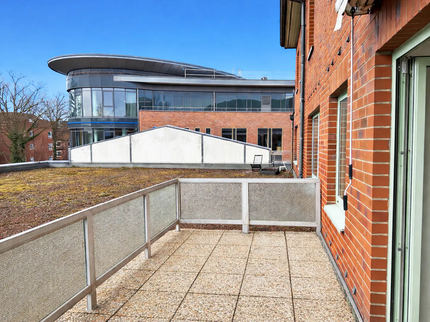 Tiled patio with a glass railing, adjacent to a red brick building. In the background is a modern building with a curved roof.