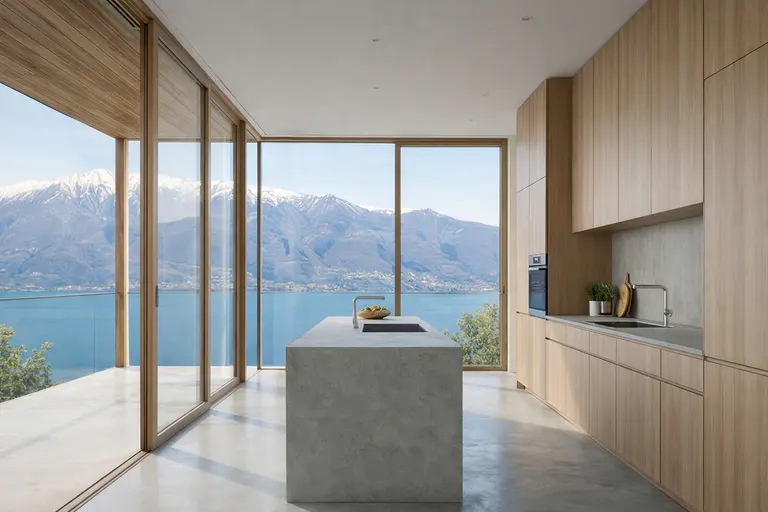 Modern kitchen with light wood cabinets, concrete island, and floor-to-ceiling windows overlooking a lake and mountains.