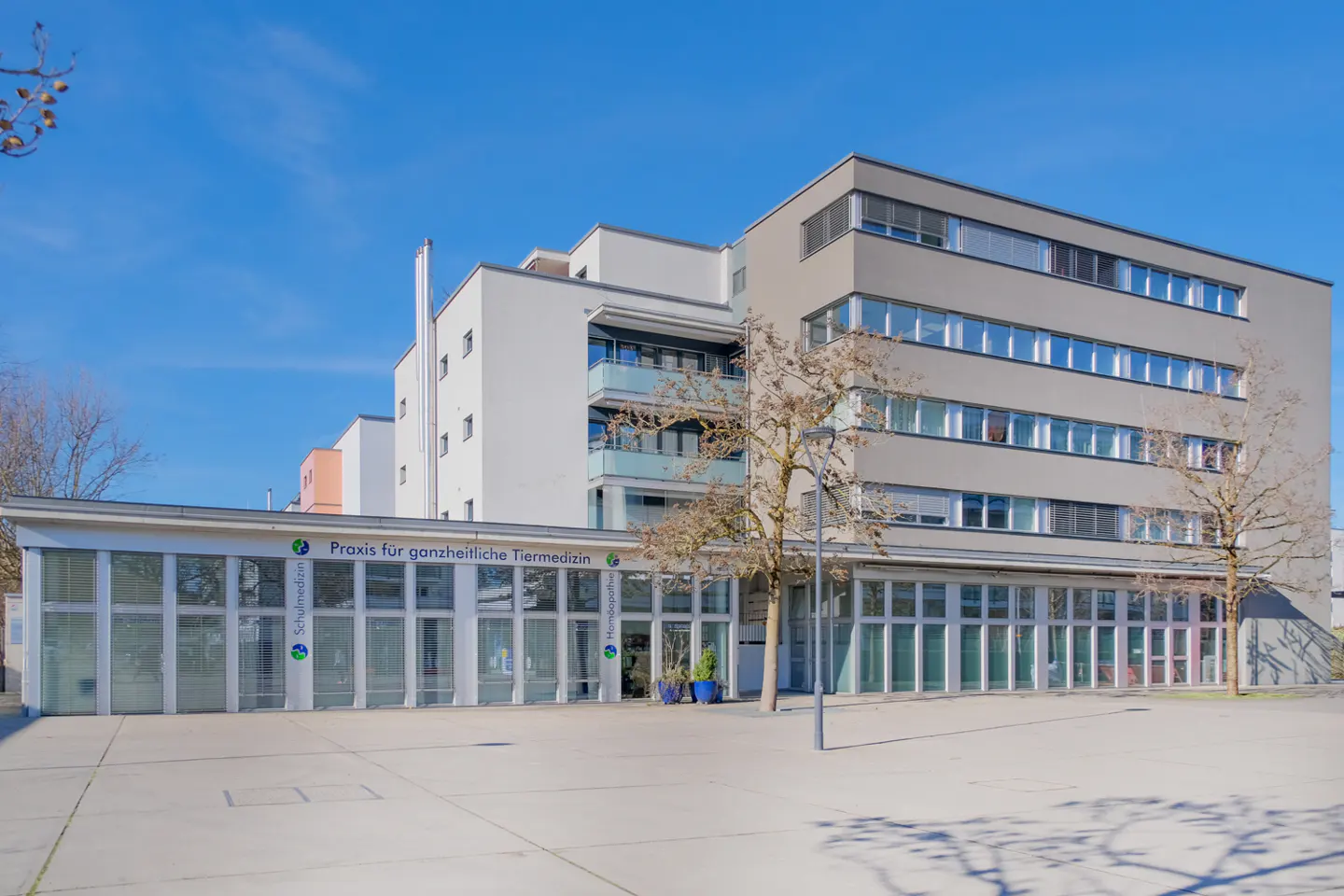 Modern building with "Praxis für ganzheitliche Tiermedizin" sign, indicating a veterinary practice. The building has a mix of white and gray sections.