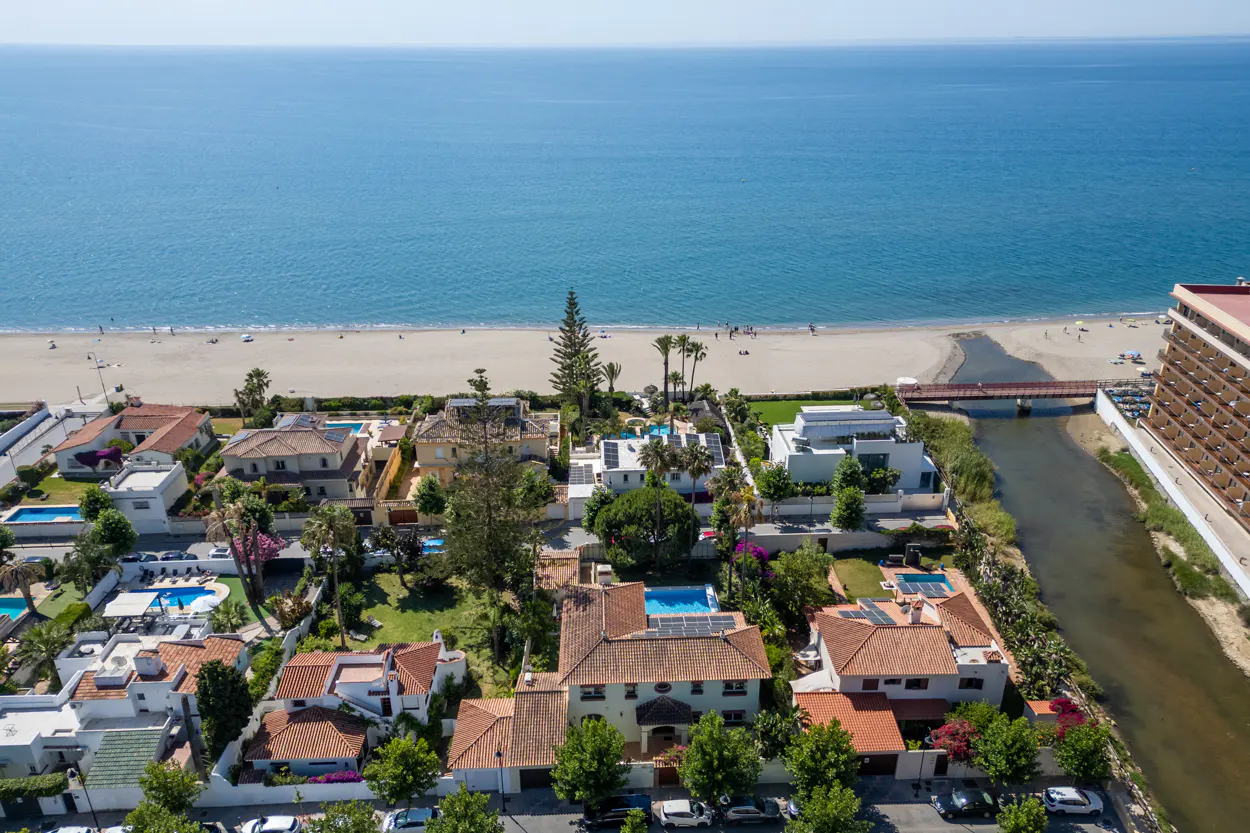 Aerial view of beachfront villas with pools, lush gardens, and a river flowing into the sea near a sandy beach.