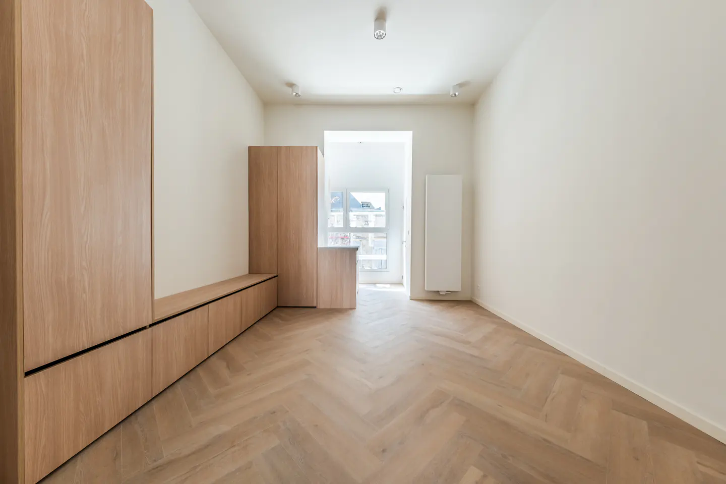 A bright, empty room with herringbone wood floors, light wood built-in cabinets, and white walls. A window lets in natural light.